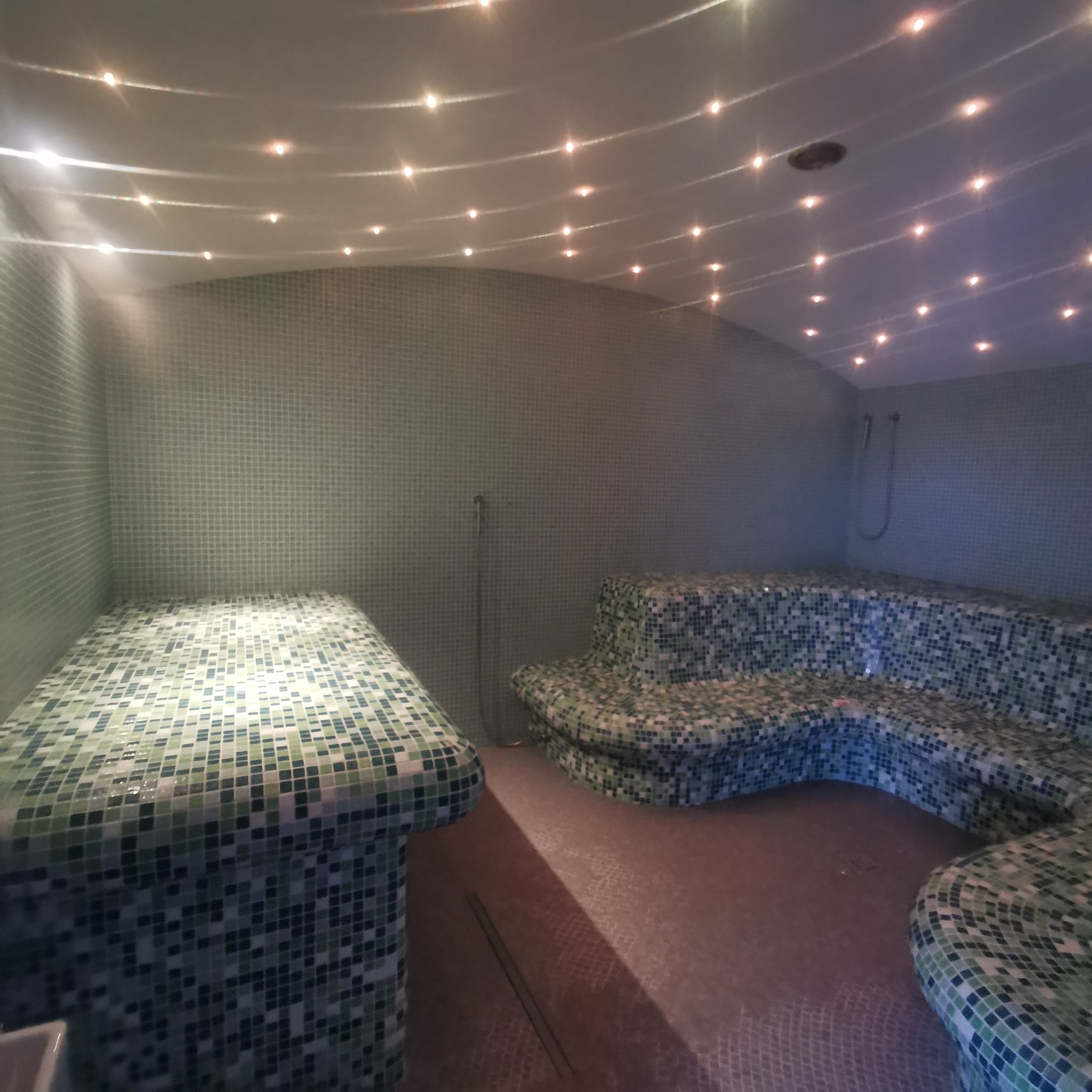 Mozaikowa łaźnia parowa z oświetleniem punktowym w suficie, wyposażona w siedziska i prysznic.