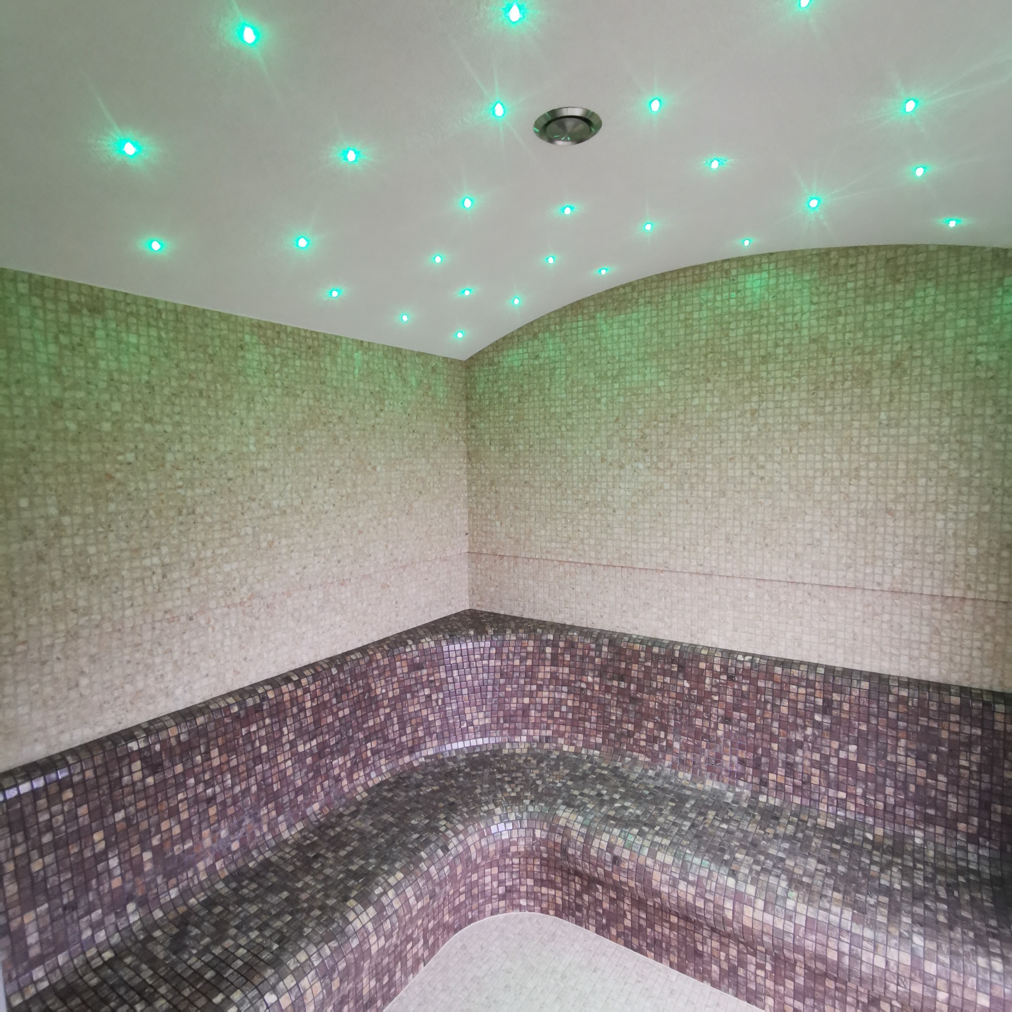 Wnętrze sauny wyłożone mozaiką w odcieniach beżu i brązu, z ławką o zaokrąglonym kształcie, oświetlone zielonymi punktowymi lampami w suficie.