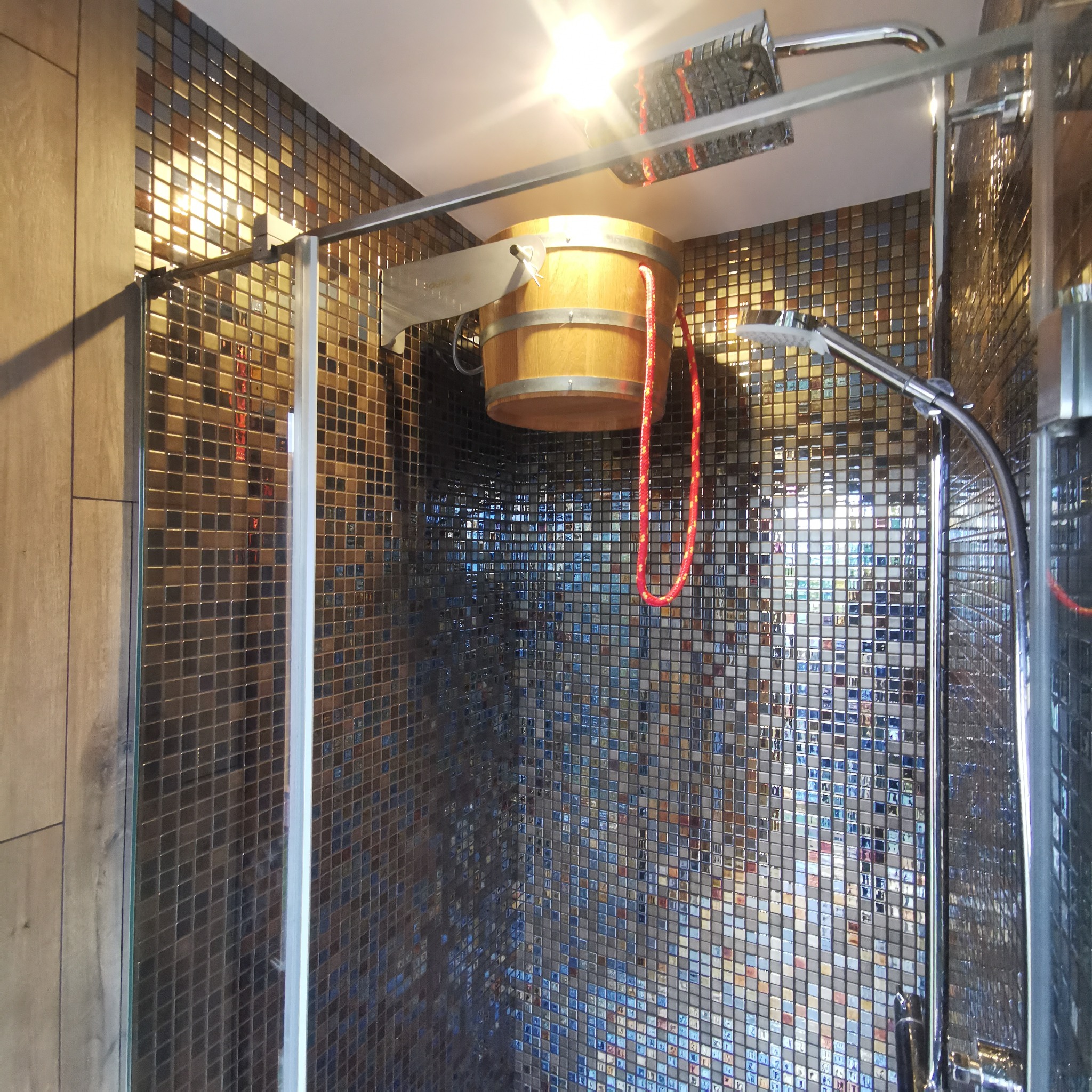 Nowoczesny prysznic z mozaiką w odcieniach brązu i niebieskiego, wyposażony w drewniany ceber z czerwonym sznurem do polewania wodą.