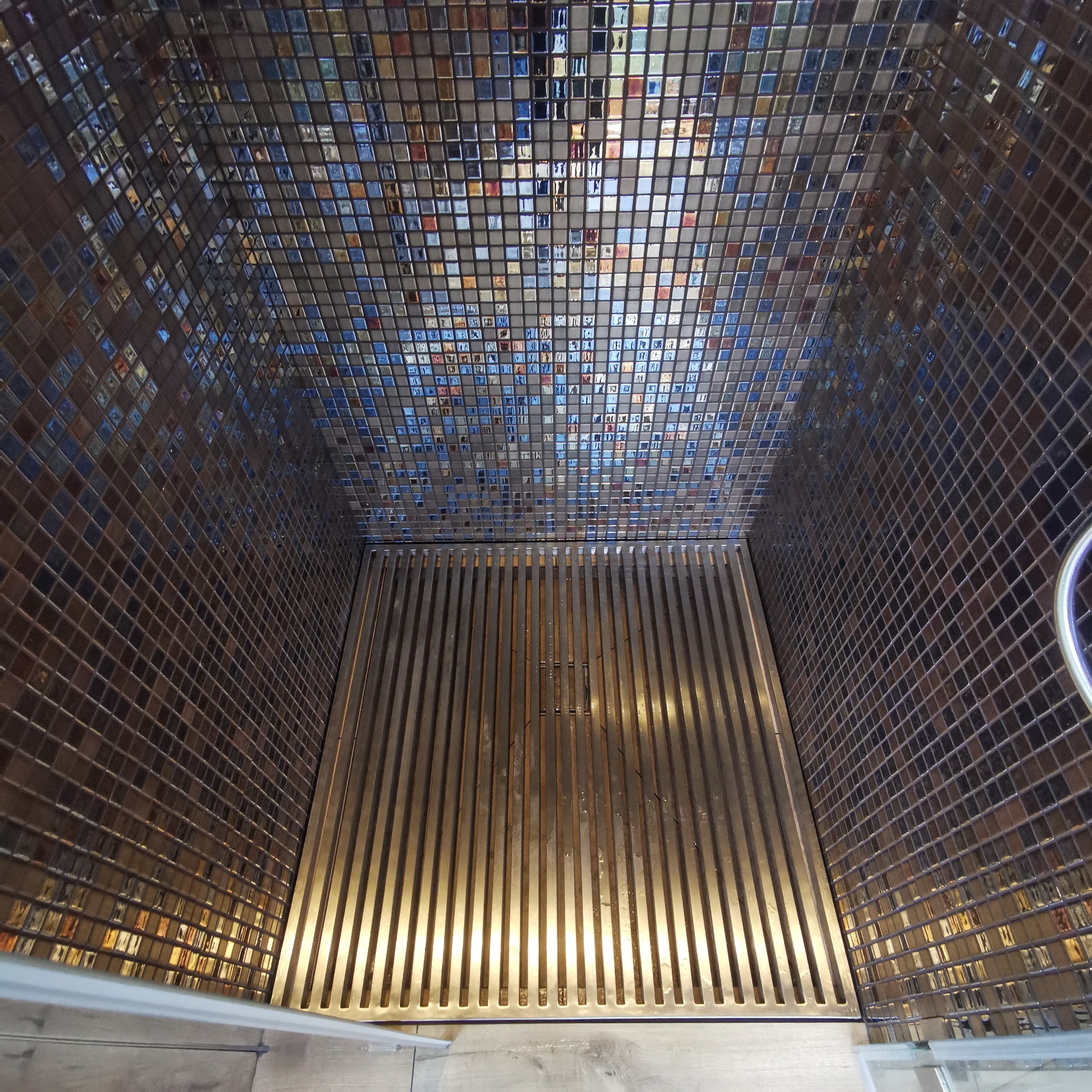 Wnętrze kabiny prysznicowej wyłożonej mozaiką z metalowym odpływem liniowym.