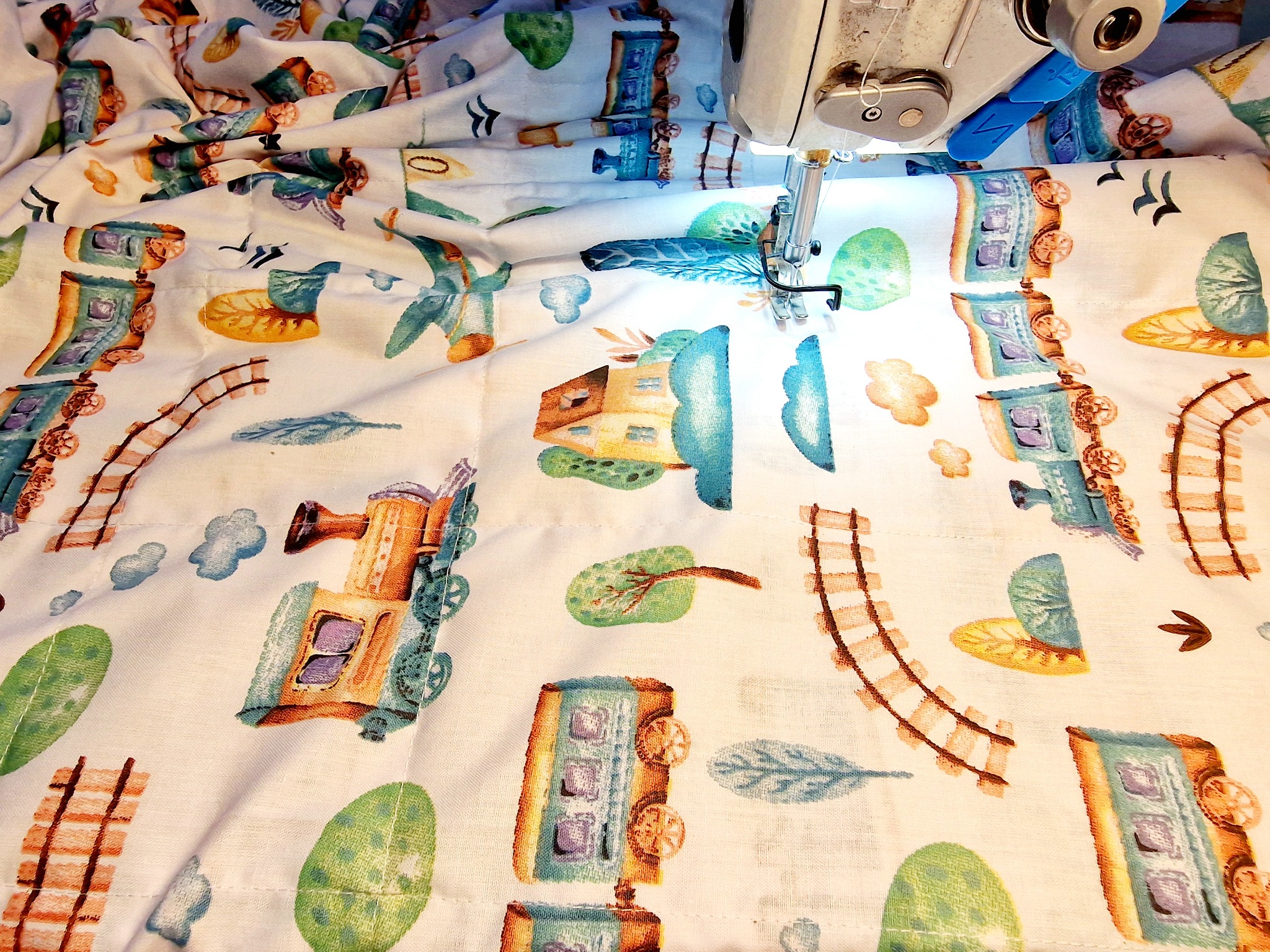 Maszyna do szycia przeszywa białą tkaninę w dziecięce wzory pociągów, torów i drzew w pastelowych kolorach.