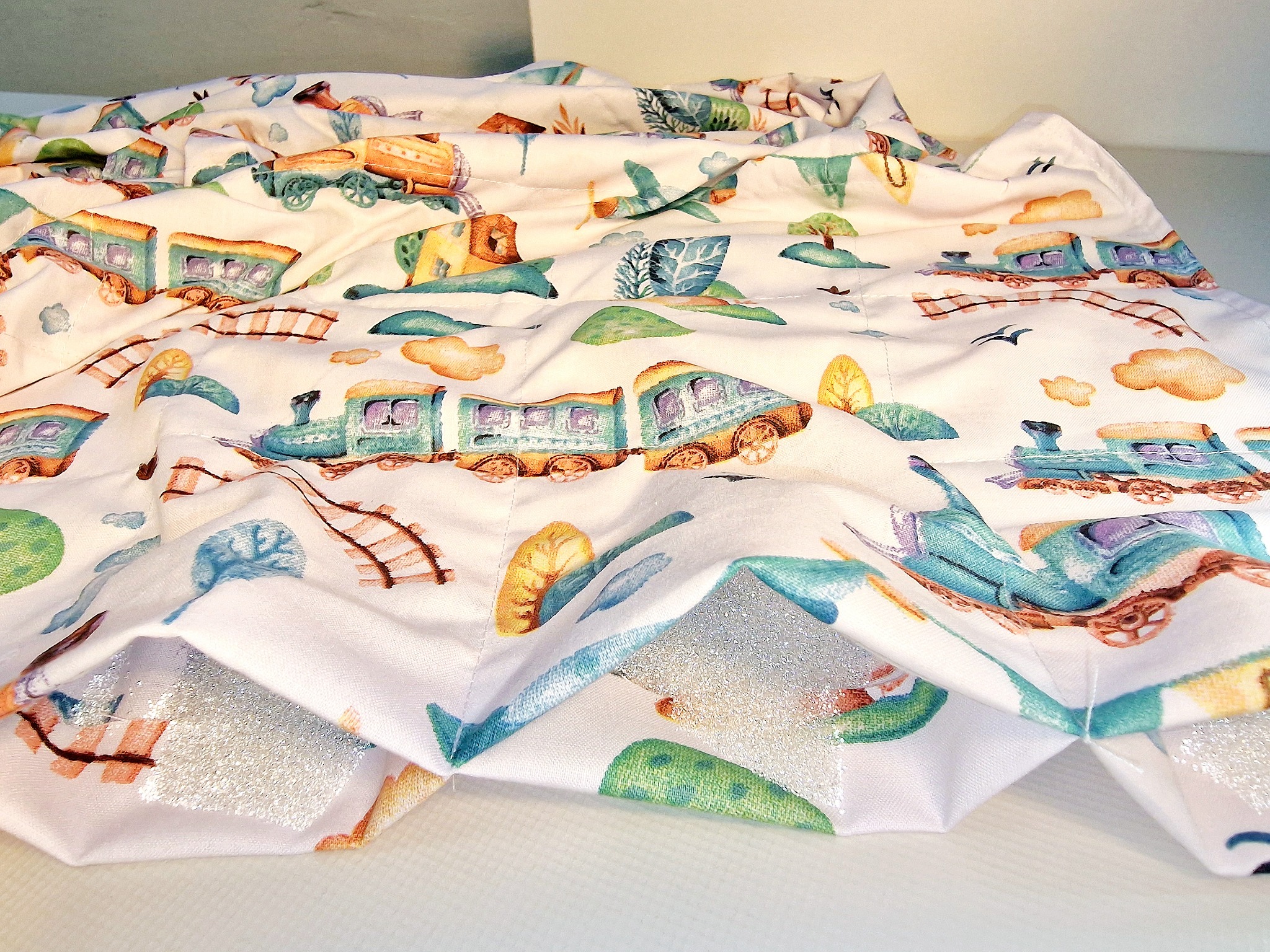 Złożona pościel dziecięca z wzorem akwarelowych pociągów, samolotów i krajobrazów, wykończona srebrną, połyskującą lamówką.