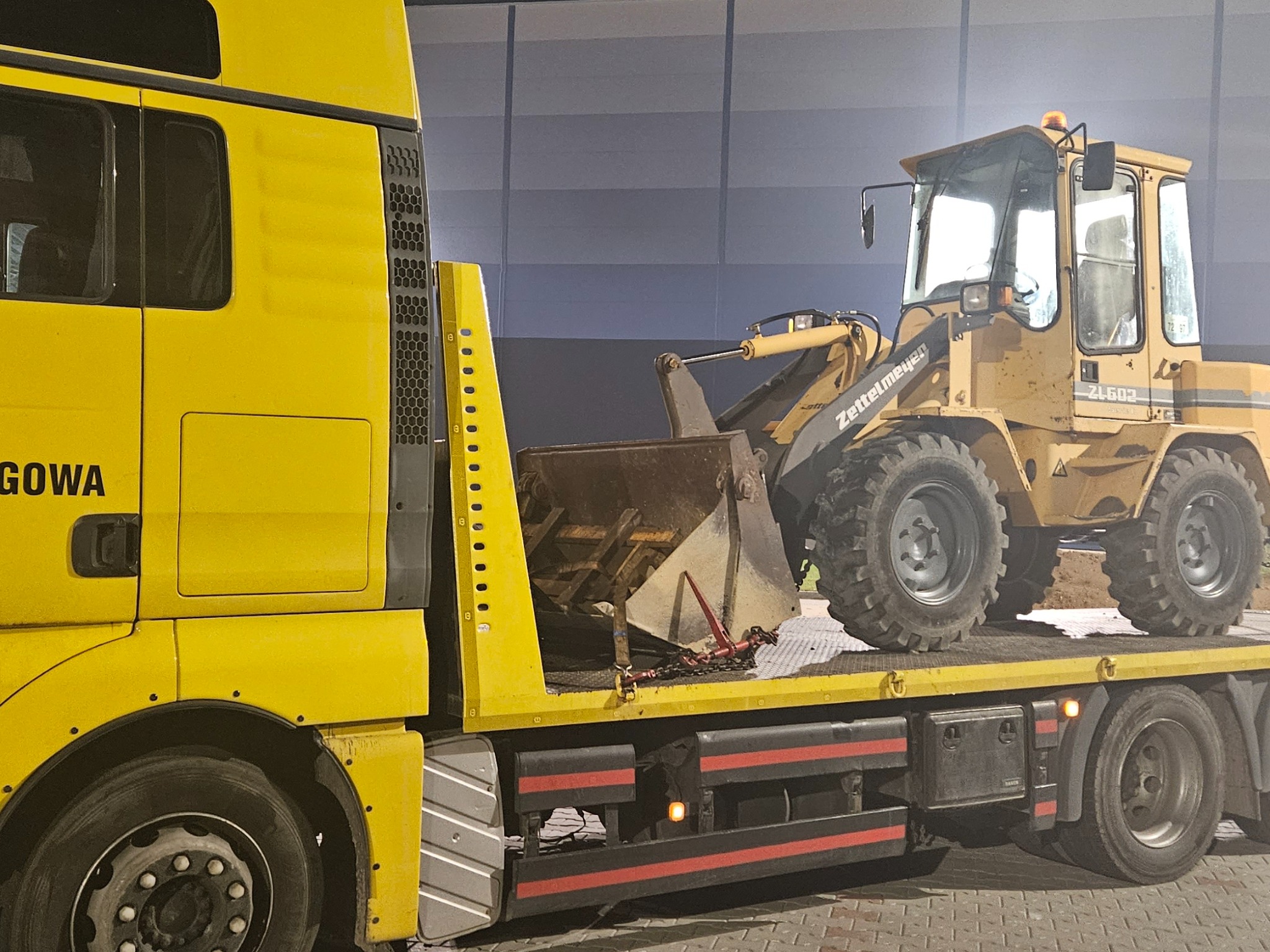Żółta ładowarka Zettelmeyer ZL602, zabezpieczona łańcuchami, przewożona na platformie żółtej ciężarówki, widok z boku.