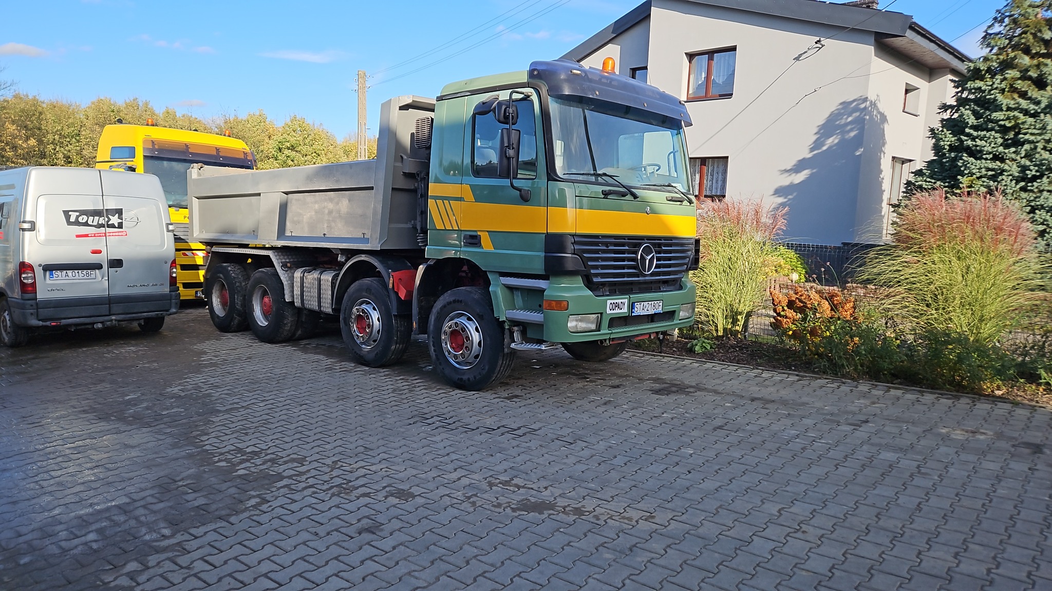 Zielono-żółta ciężarówka wywrotka Mercedes-Benz z szarą skrzynią ładunkową, zaparkowana na szarej kostce brukowej, z widoczną tablicą rejestracyjną i oznaczeniem 'ODPADY', obok zaparkowany biały...