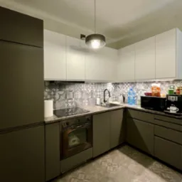 Nowoczesna kuchnia z szarymi szafkami, białymi szafkami wiszącymi, wzorzystymi płytkami nad blatem i na podłodze, oświetlona wiszącą lampą.