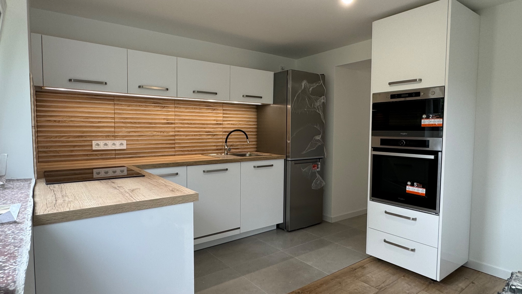 Nowoczesna kuchnia z białymi szafkami, drewnianym blatem i panelem ściennym, srebrną lodówką w folii ochronnej oraz zabudowanym piekarnikiem z naklejkami informacyjnymi.