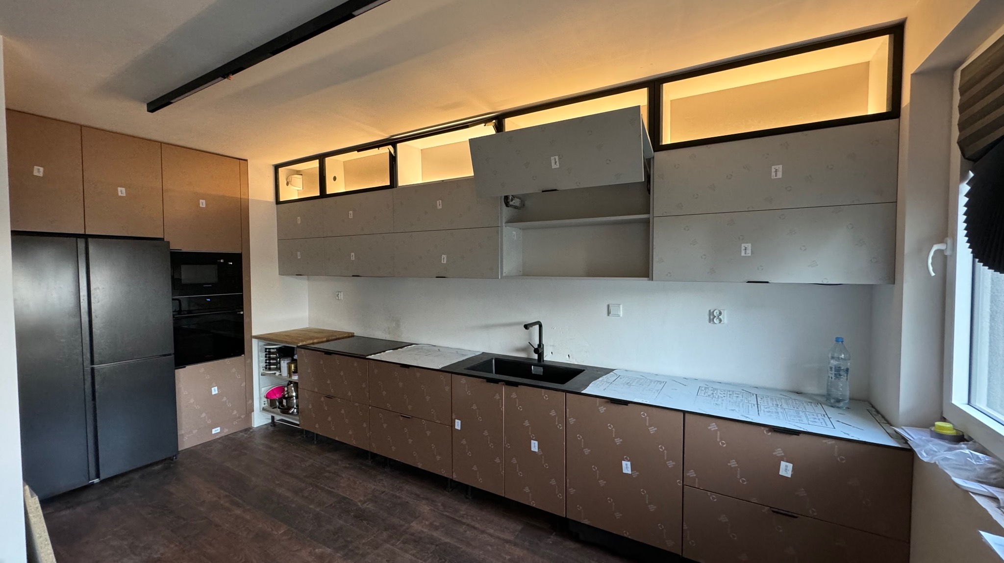 Nowoczesna kuchnia w trakcie montażu: dolne szafki z brązowymi frontami zabezpieczone folią, górne szafki z podświetleniem, otwarte drzwiczki, widoczna czarna lodówka i piekarnik w zabudowie...
