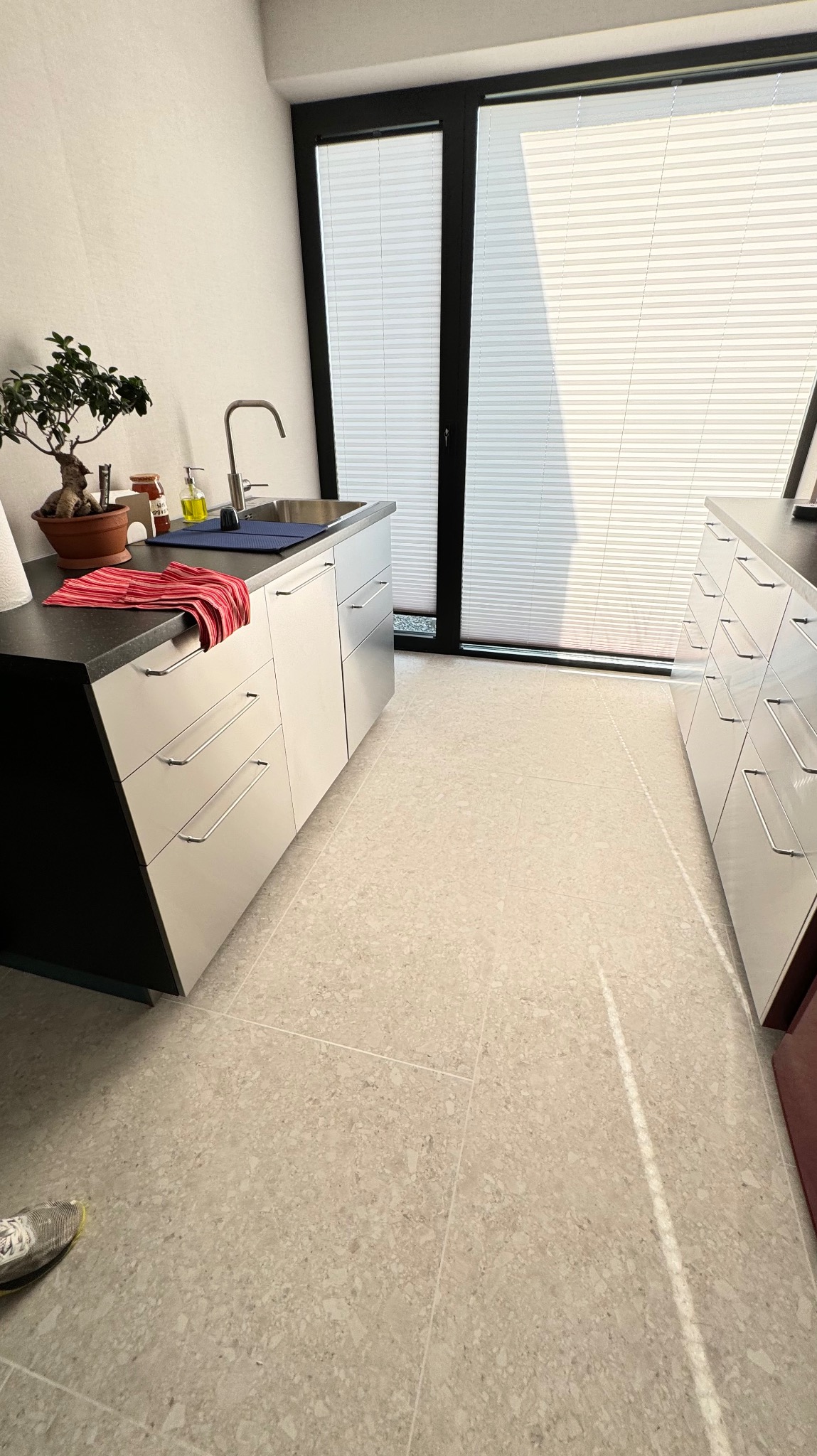 Nowoczesna kuchnia z białymi szafkami o minimalistycznym designie, czarnym blatem i dużym oknem z żaluzjami, uzupełniona o bonsai i tekstylia w kolorze czerwonym. Widoczny fragment buta w lewym...