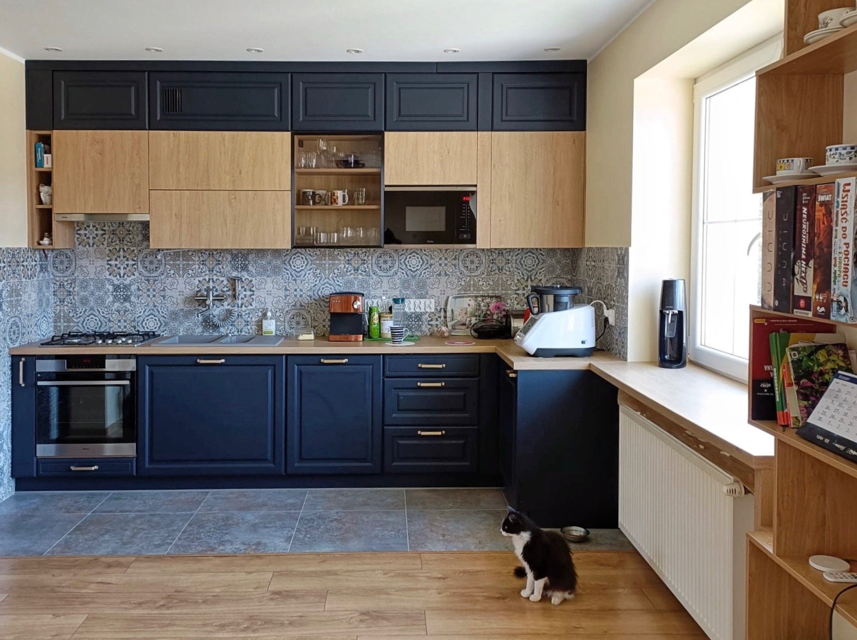 Nowoczesna kuchnia z ciemnymi i drewnianymi szafkami, ozdobnymi płytkami na ścianie i czarno-białym kotem siedzącym na podłodze