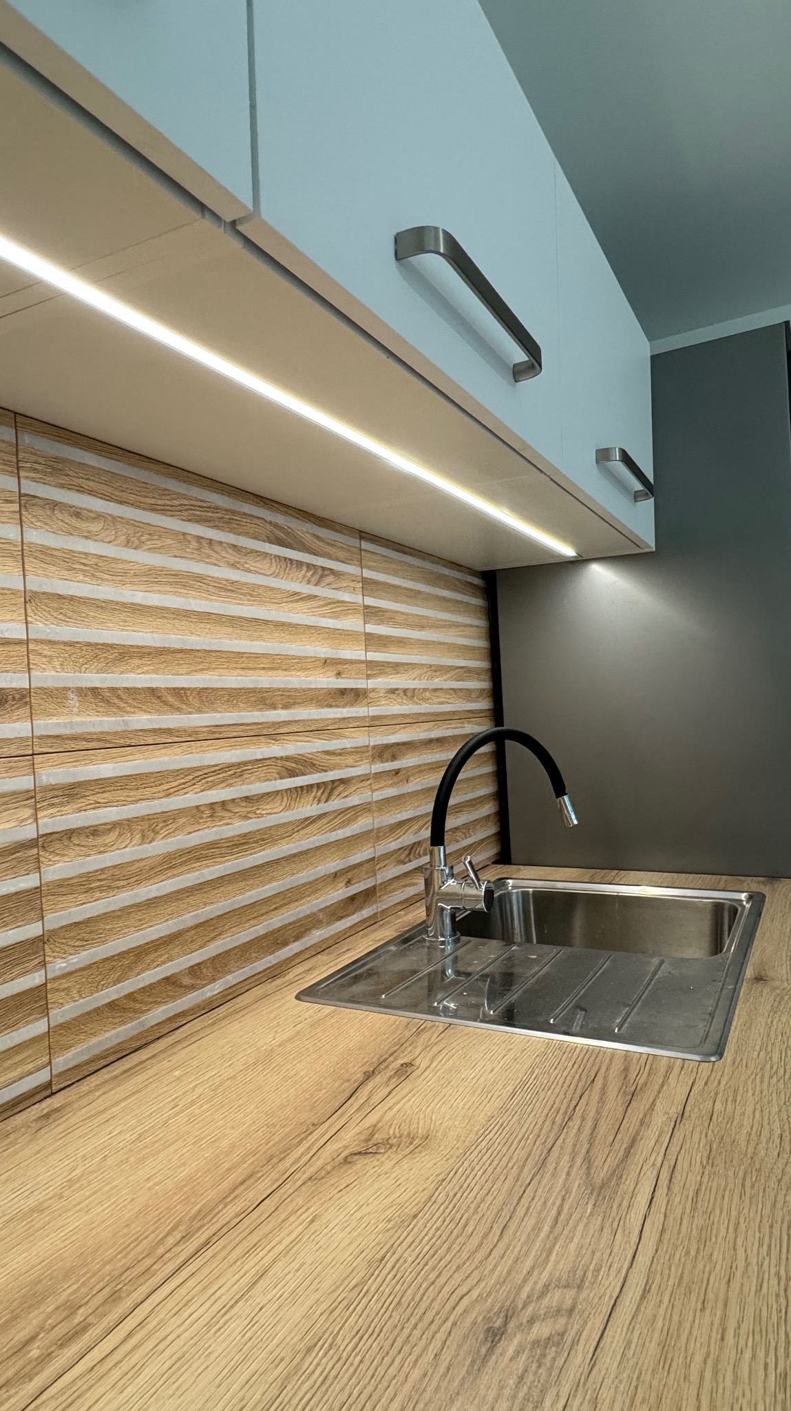 Nowoczesna kuchnia z drewnianym blatem i ozdobną ścianą z płytek w paski, oświetlenie LED pod szafkami, metalowy zlew i czarna bateria.