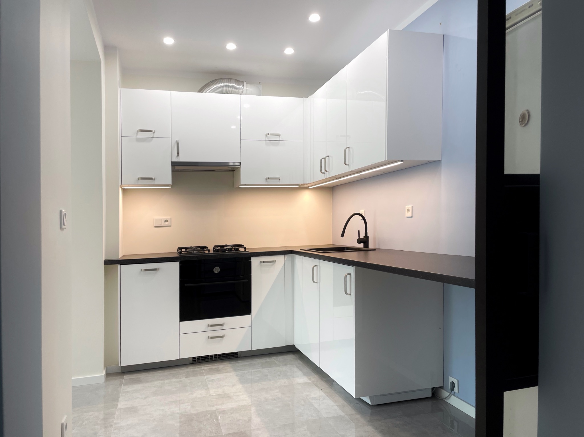 Nowoczesna, biała kuchnia z czarnym blatem i armaturą, oświetlona punktowymi lampami sufitowymi i listwą LED pod szafkami wiszącymi; widoczny fragment srebrnego przewodu wentylacyjnego.