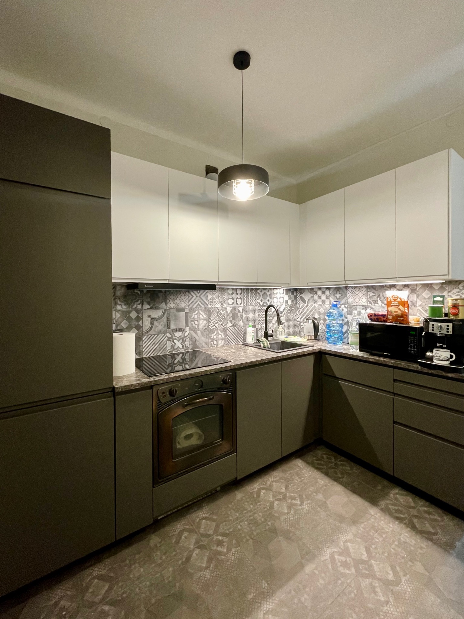 Nowoczesna kuchnia z szarymi szafkami, białymi szafkami wiszącymi, wzorzystymi płytkami nad blatem i na podłodze, oświetlona wiszącą lampą.