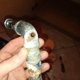 Hydro4 - Zardzewiały, skorodowany zawór hydrauliczny z zatkanym otworem, trzymany w dłoni na tle płytek podłogowych.