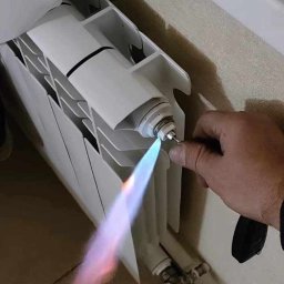 Hydro4 - Biały grzejnik panelowy, lutowanie miedzianej rurki palnikiem gazowym, widoczny płomień i ręka montera.