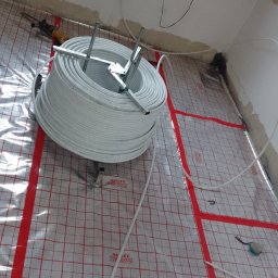 Hydro4 - Rozwinięta rolka rur do instalacji podłogowej na folii izolacyjnej z czerwoną siatką, widoczne narzędzia i surowy stan wykończenia pomieszczenia.