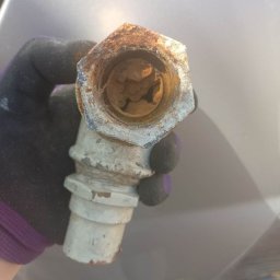 Hydro4 - Zardzewiała złączka hydrauliczna, trzymana w rękawicy roboczej, widoczne osady wewnątrz.