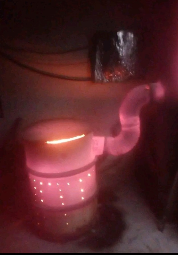 Niedoskonałe zdjęcie pieca typu 'koza' z rurą dymową, oświetlonego czerwonym światłem, widoczne otwory wentylacyjne w korpusie.