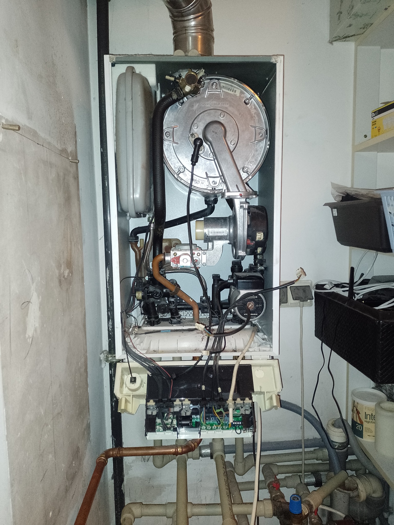 Otwarty piec gazowy Viessmann z widocznymi elementami wewnętrznymi, rurami miedzianymi i plastikowymi, instalacją elektryczną, na tle ściany i półki z akcesoriami.