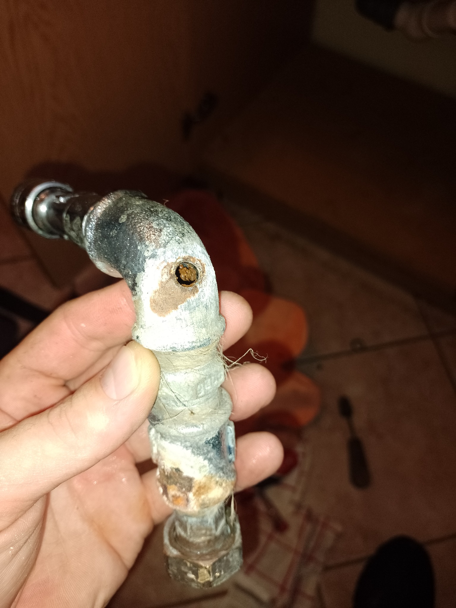 Zardzewiały, skorodowany zawór hydrauliczny z zatkanym otworem, trzymany w dłoni na tle płytek podłogowych.