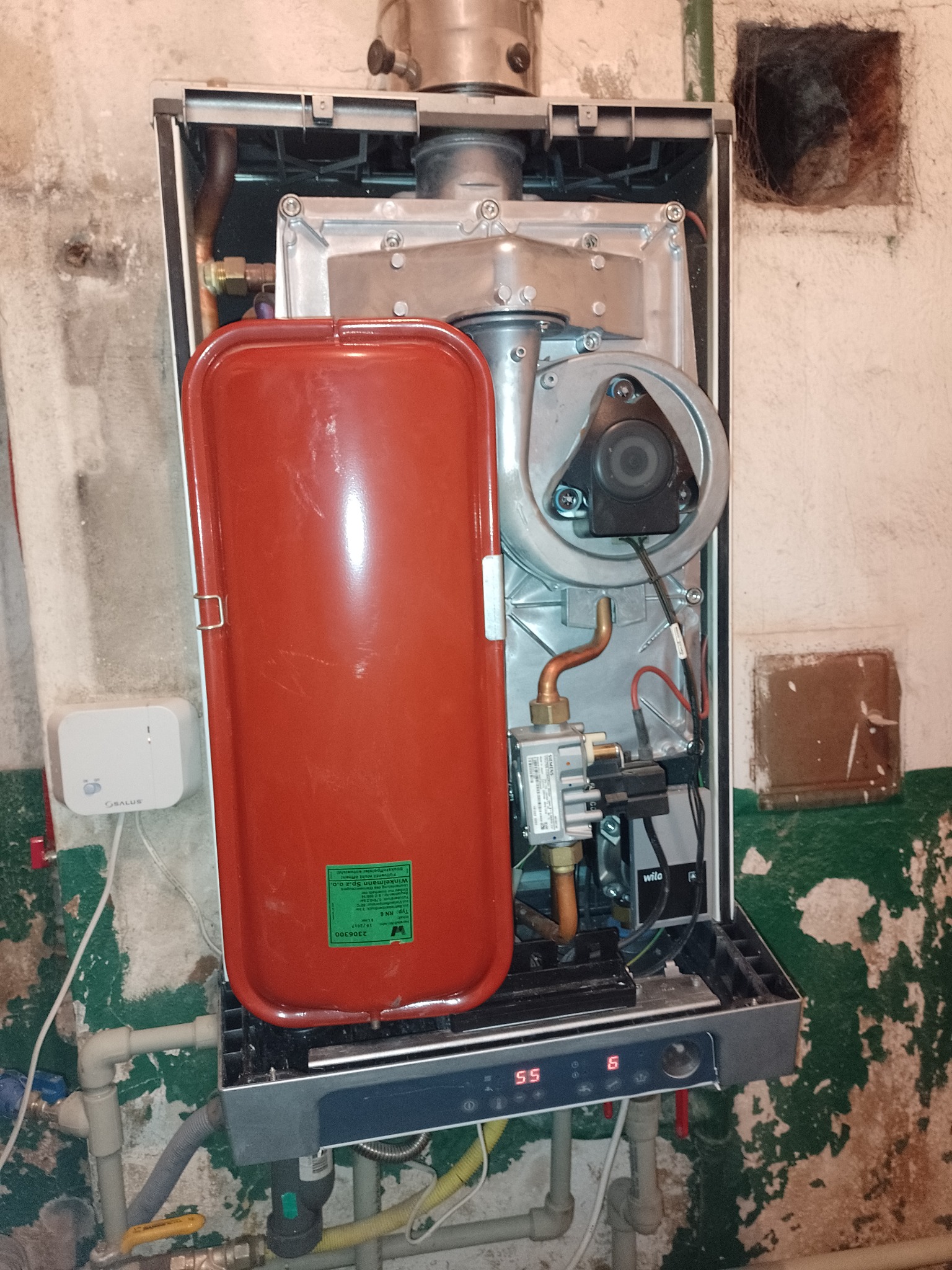 Otwarty piec gazowy z widocznymi elementami wewnętrznymi, czerwonym zbiornikiem wyrównawczym i podłączeniami hydraulicznymi na tle ściany ze śladami farby.