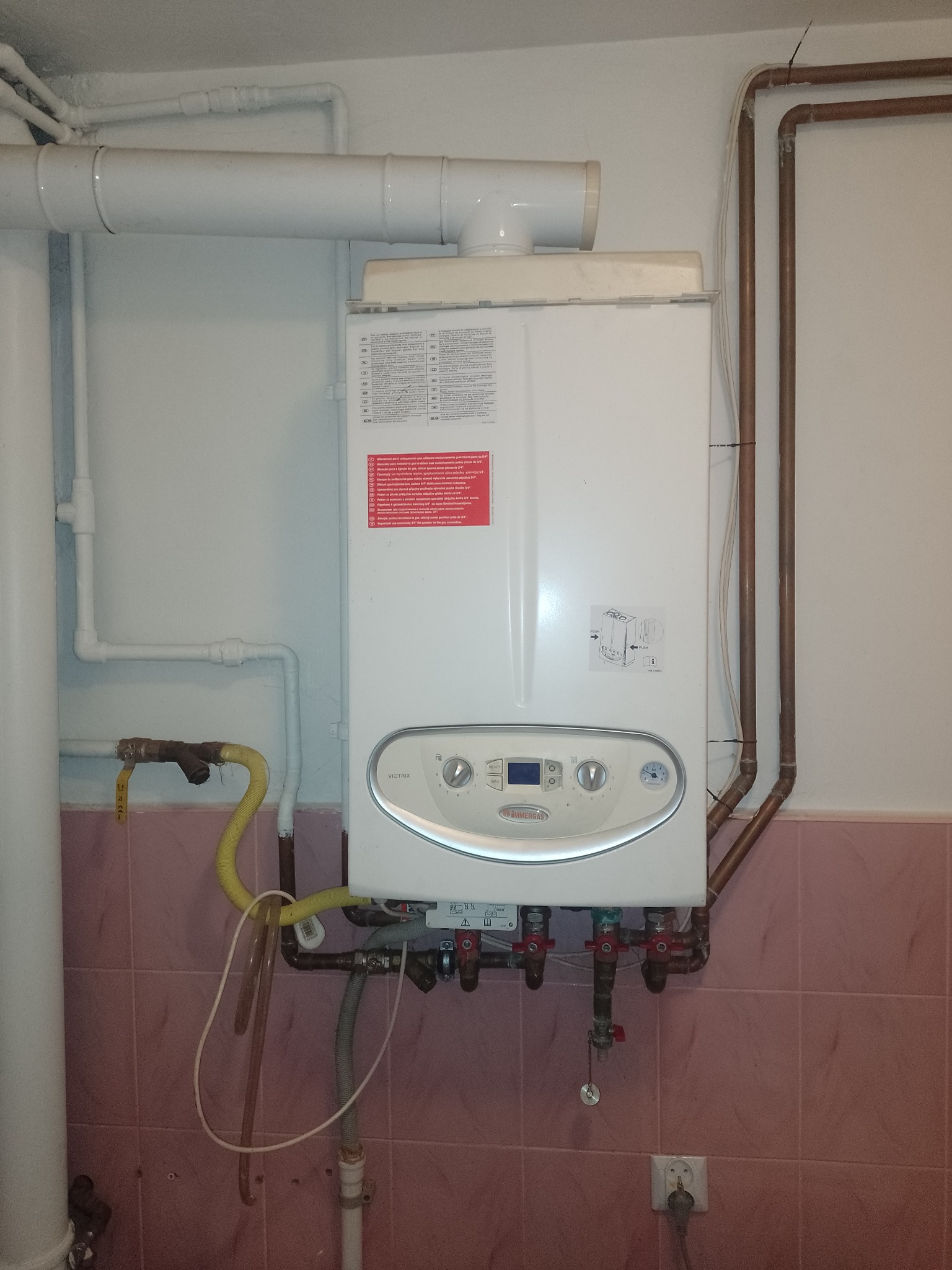 Biały piec gazowy Immergas Victrix z podłączonymi rurami miedzianymi i plastikowymi, zamontowany na różowej ścianie z płytek, widoczne zawory i przewody.