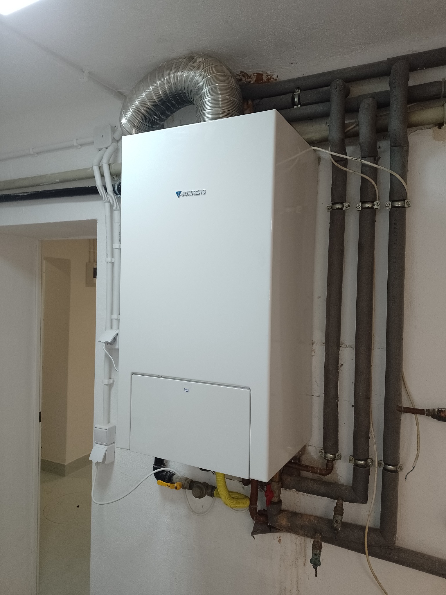 Nowy, biały piec gazowy Junkers zamontowany na ścianie z widocznymi rurami i przewodami, podłączony do komina wentylacyjnego.