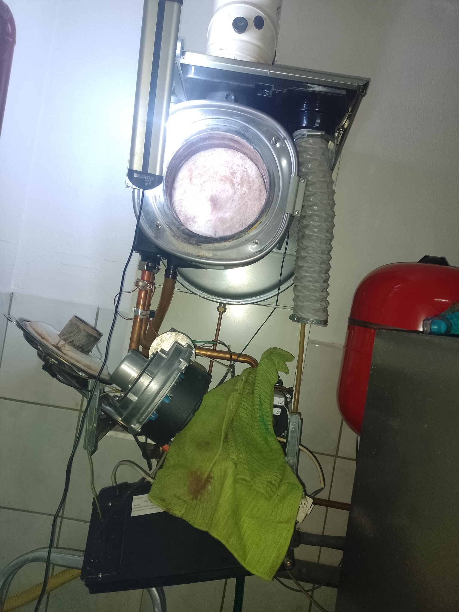 Otwarty piec gazowy w trakcie serwisu, widoczne elementy wewnętrzne, zielona ściereczka i instalacja z miedzianych rur.