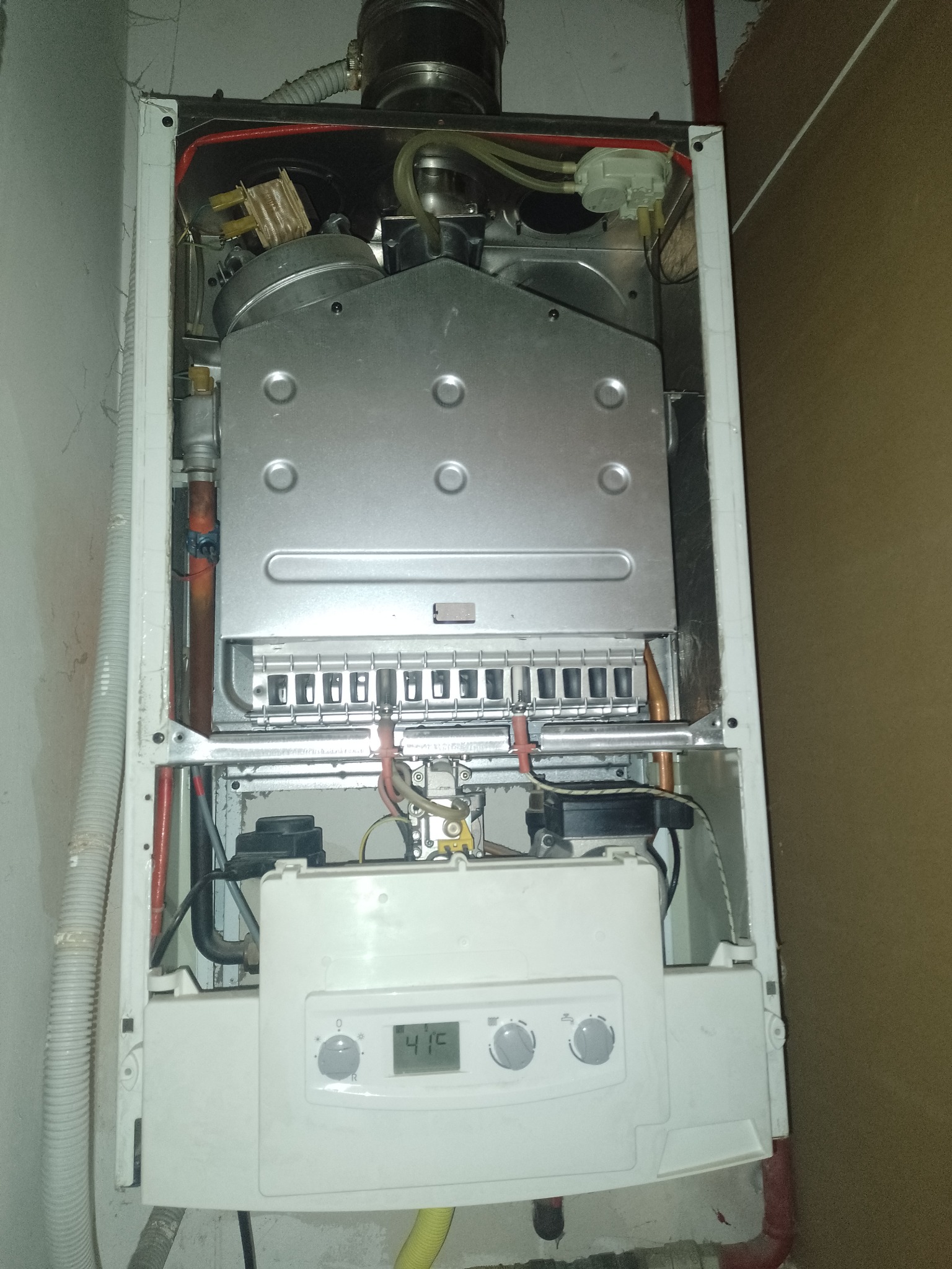 Otwarty piec gazowy z widocznymi elementami wewnętrznymi, w tym srebrną obudową, przewodami i panelem sterowania z wyświetlaczem wskazującym temperaturę, zamontowany na ścianie z widocznymi śladami...