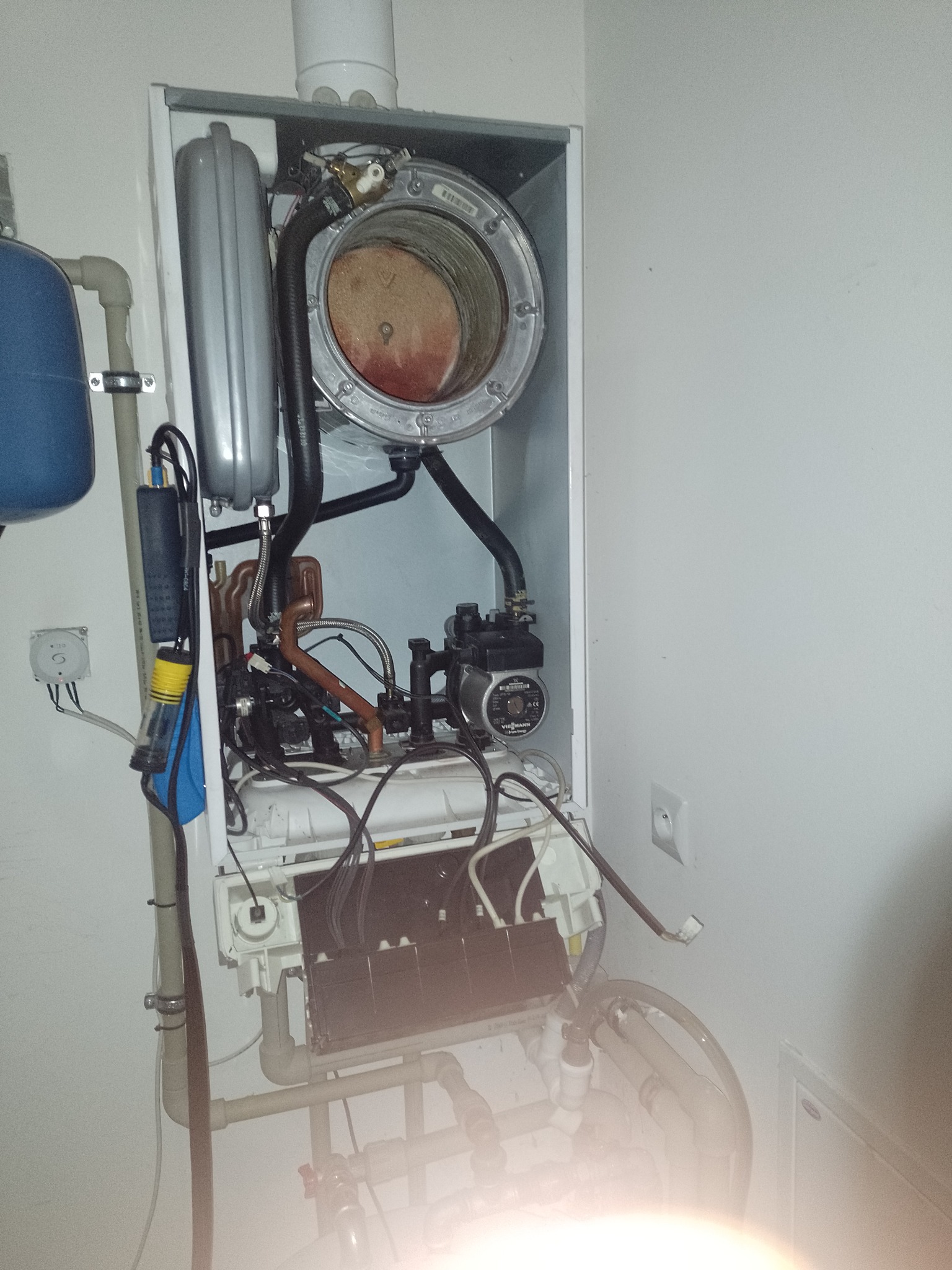 Otwarty piec gazowy w trakcie serwisu, widoczne elementy wewnętrzne, rury, przewody i pompa obiegowa, ślady użytkowania na palniku.