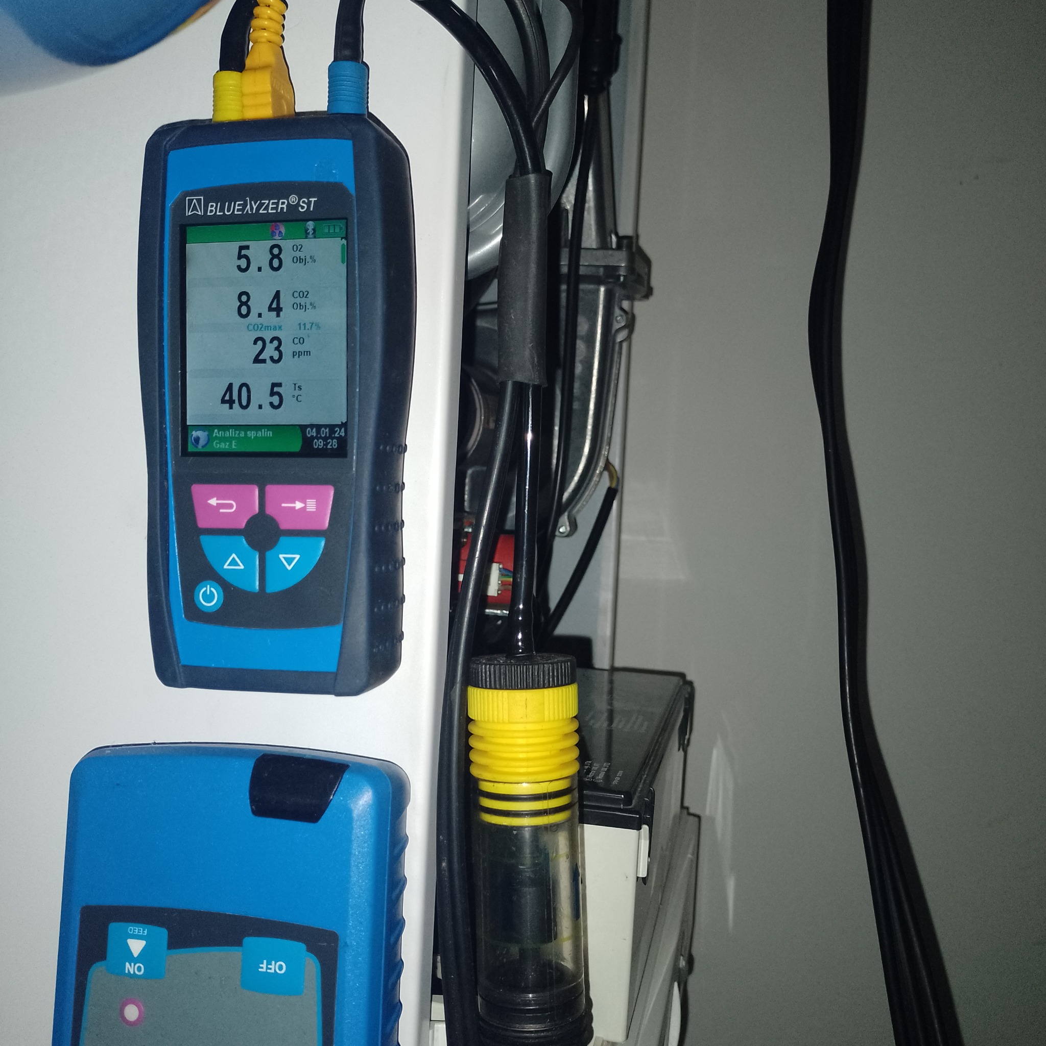 Analizator spalin BlueLyzer ST z wyświetlanymi wartościami O2, CO2 i CO, podłączony do instalacji za pomocą sondy z żółtą końcówką. Widoczny fragment kotła gazowego i przewody.