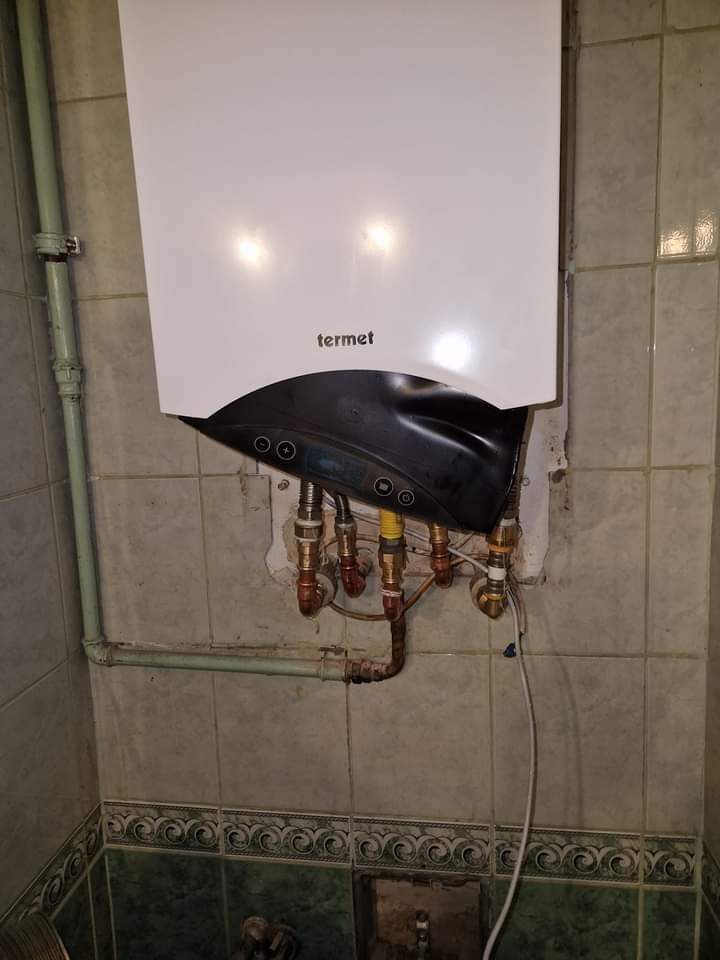 Biały piec gazowy Termet zamontowany na kafelkowanej ścianie z widocznymi rurami i instalacją hydrauliczną, w trakcie podłączenia, z naruszoną strukturą tynku.