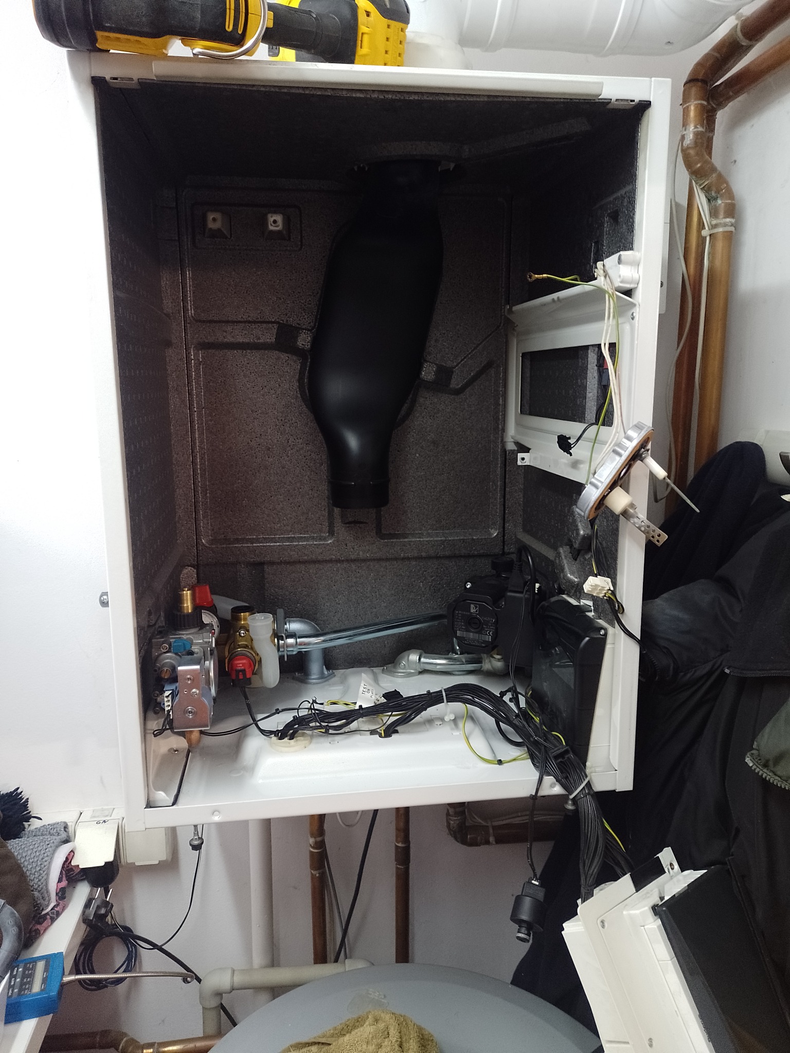 Otwarty piec gazowy w trakcie naprawy, widoczne elementy wewnętrzne, przewody i narzędzia w tle, instalacja hydrauliczna z rur miedzianych.