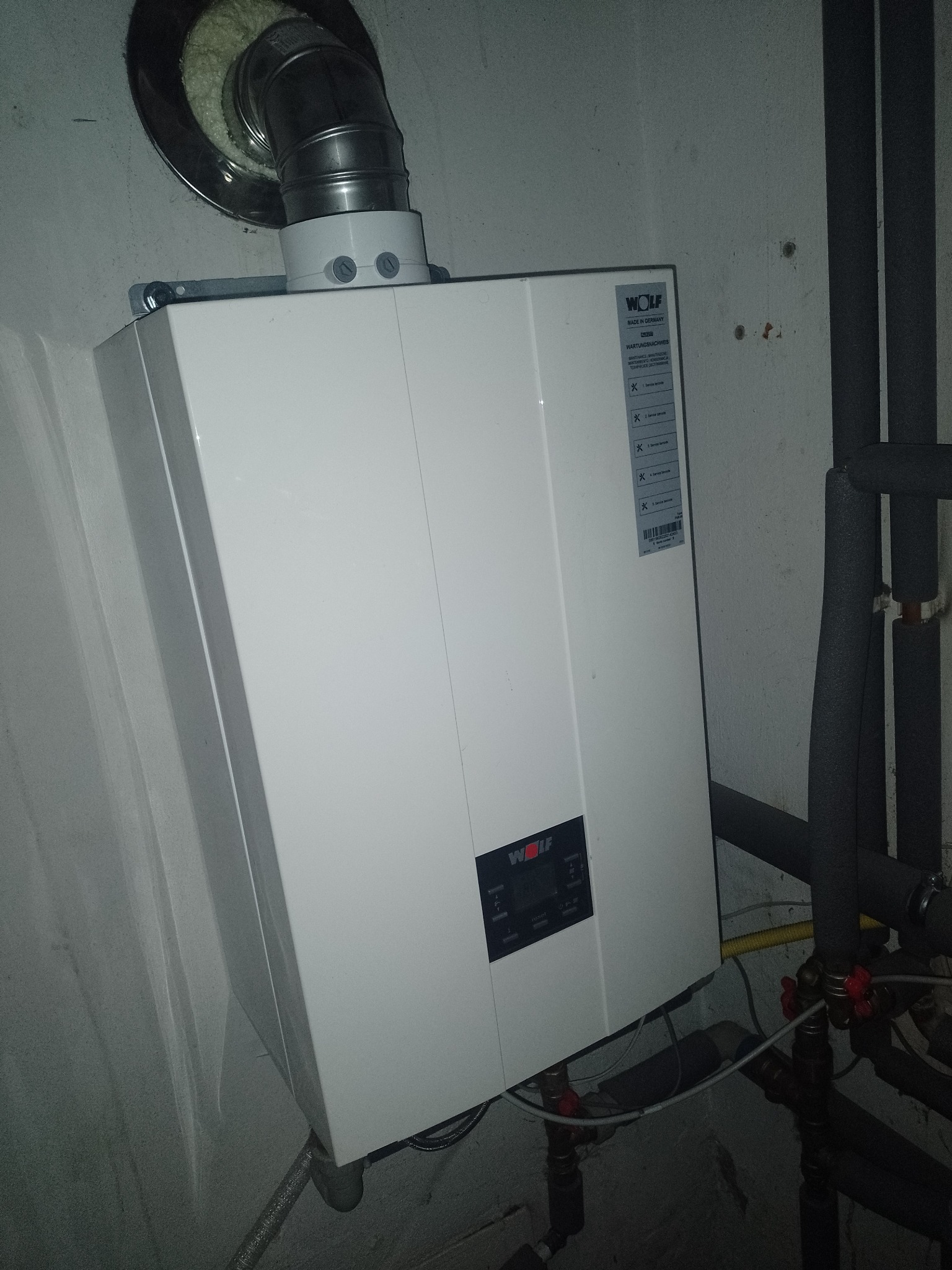 Biały piec gazowy marki Wolf z podłączonym kominem i instalacją, widoczne rury z izolacją termiczną i zawory, zamontowany na szarej ścianie w piwnicy.