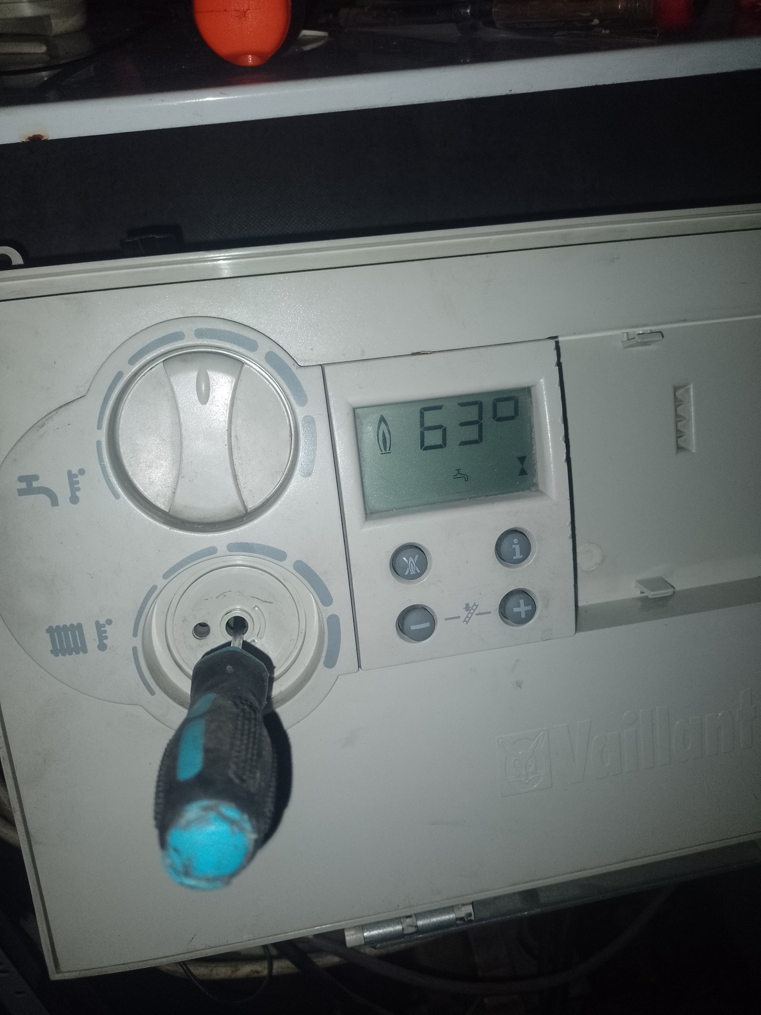 Śrubokręt z niebieską rączką w gnieździe panelu sterowania pieca gazowego Vaillant, wyświetlacz pokazuje temperaturę 63 stopnie.