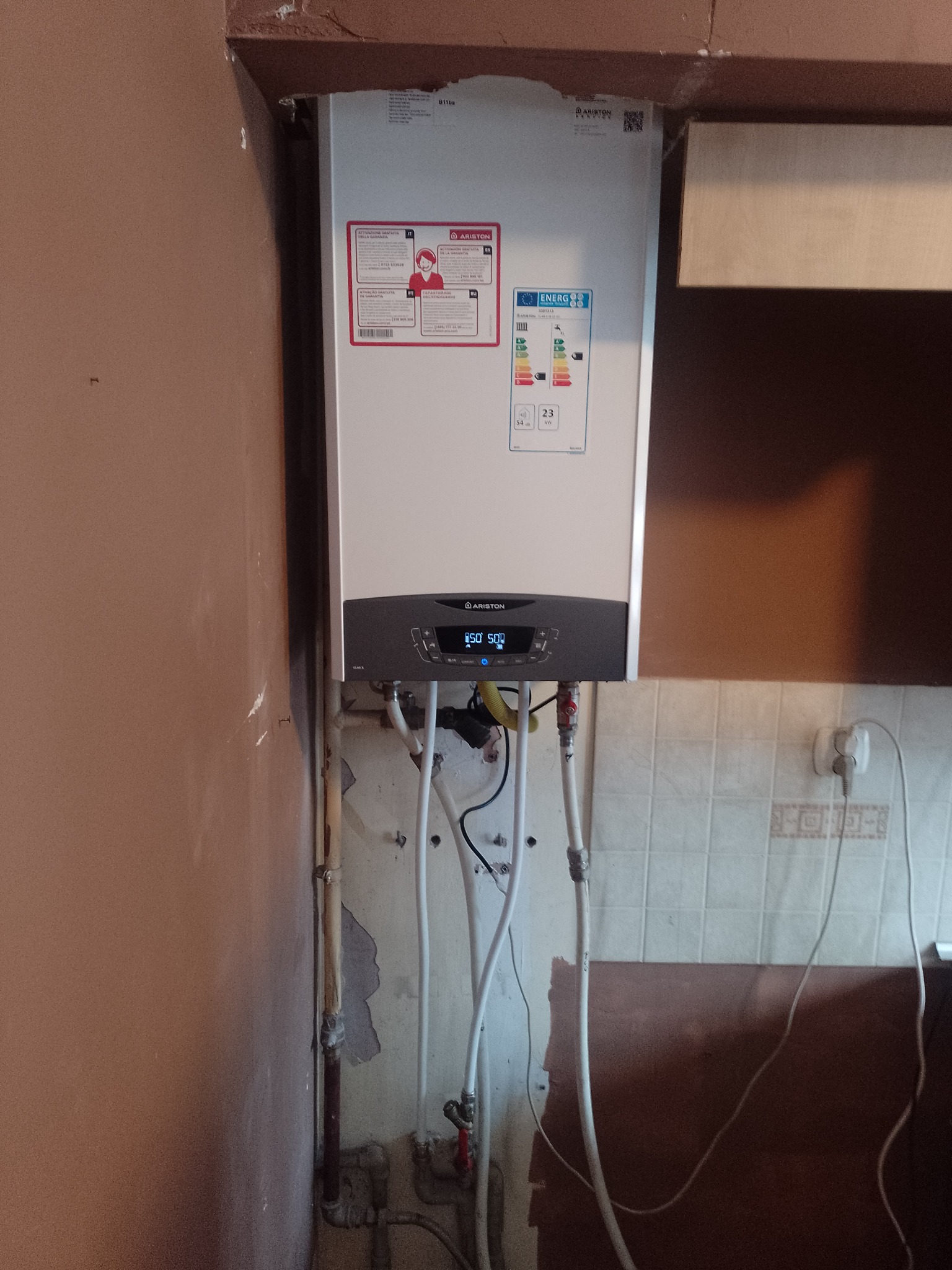 Nowy piec gazowy Ariston zamontowany na ścianie z widocznymi rurami instalacji w trakcie podłączenia, z wyświetlaczem wskazującym temperaturę.