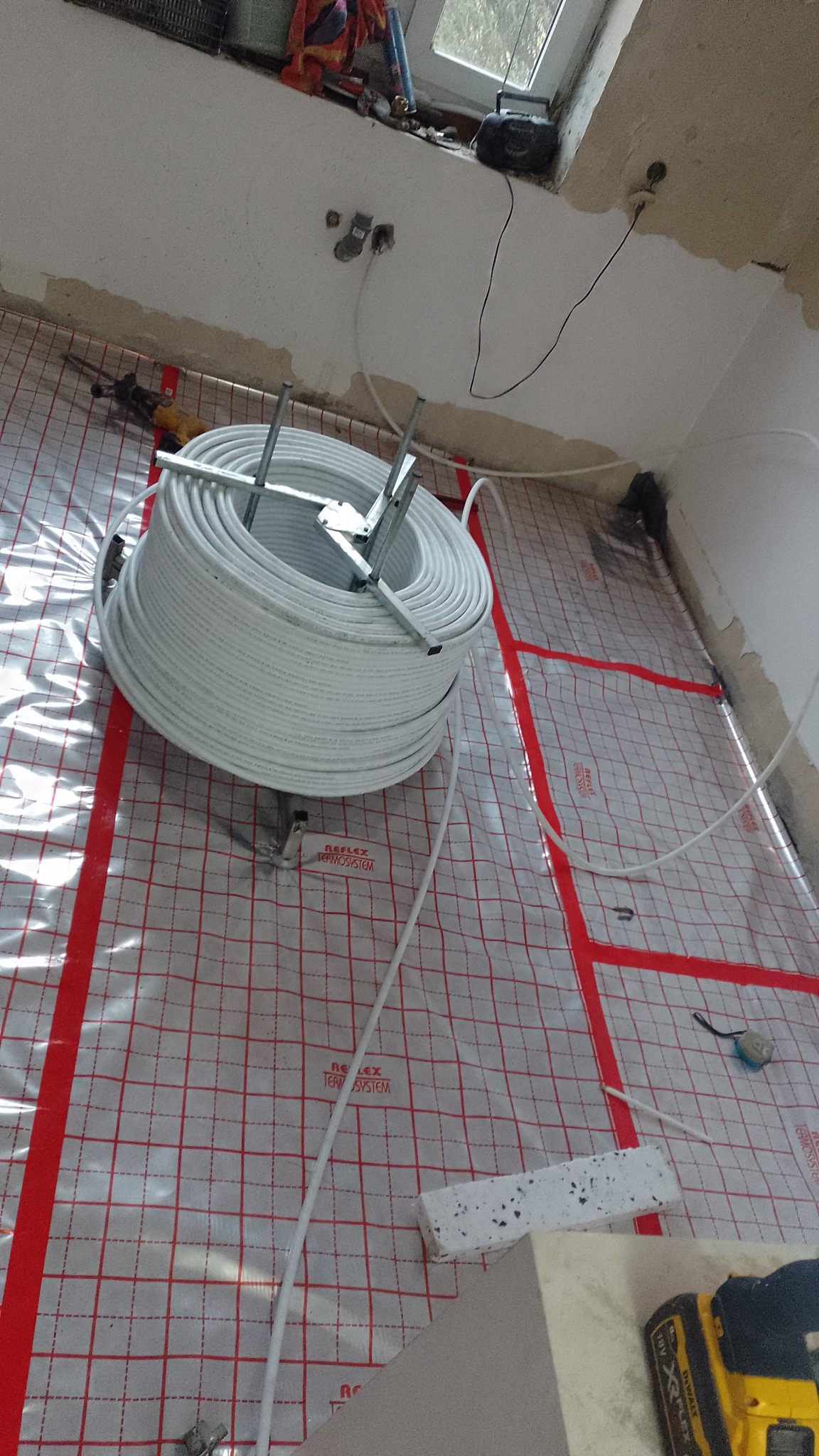 Rozwinięta rolka rur do instalacji podłogowej na folii izolacyjnej z czerwoną siatką, widoczne narzędzia i surowy stan wykończenia pomieszczenia.
