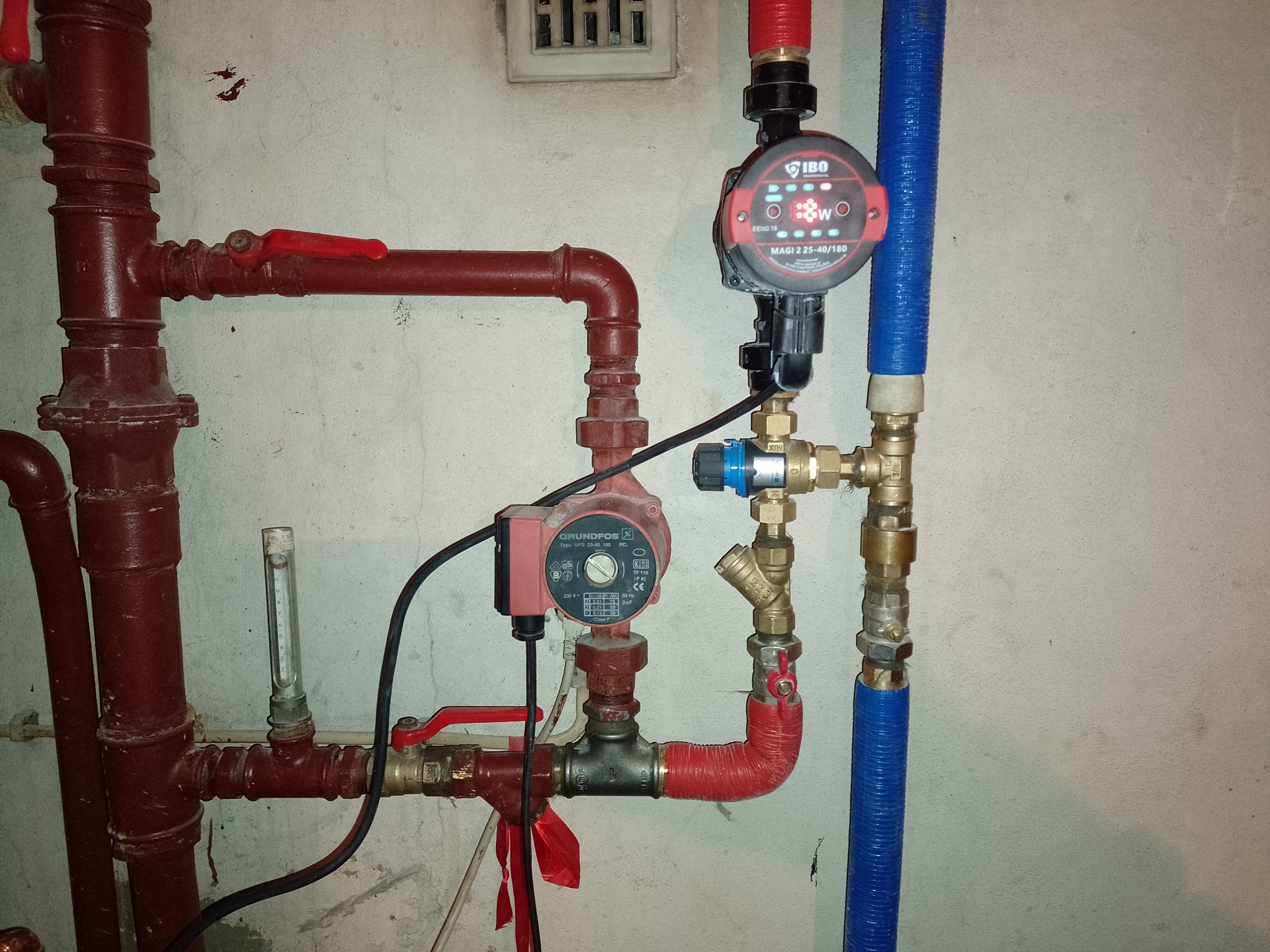 Rozbudowana instalacja hydrauliczna z dwoma pompami obiegowymi Grundfos i IBO, rury w kolorze czerwonym i niebieskim, zawory, manometr, całość zamontowana na szarej ścianie.