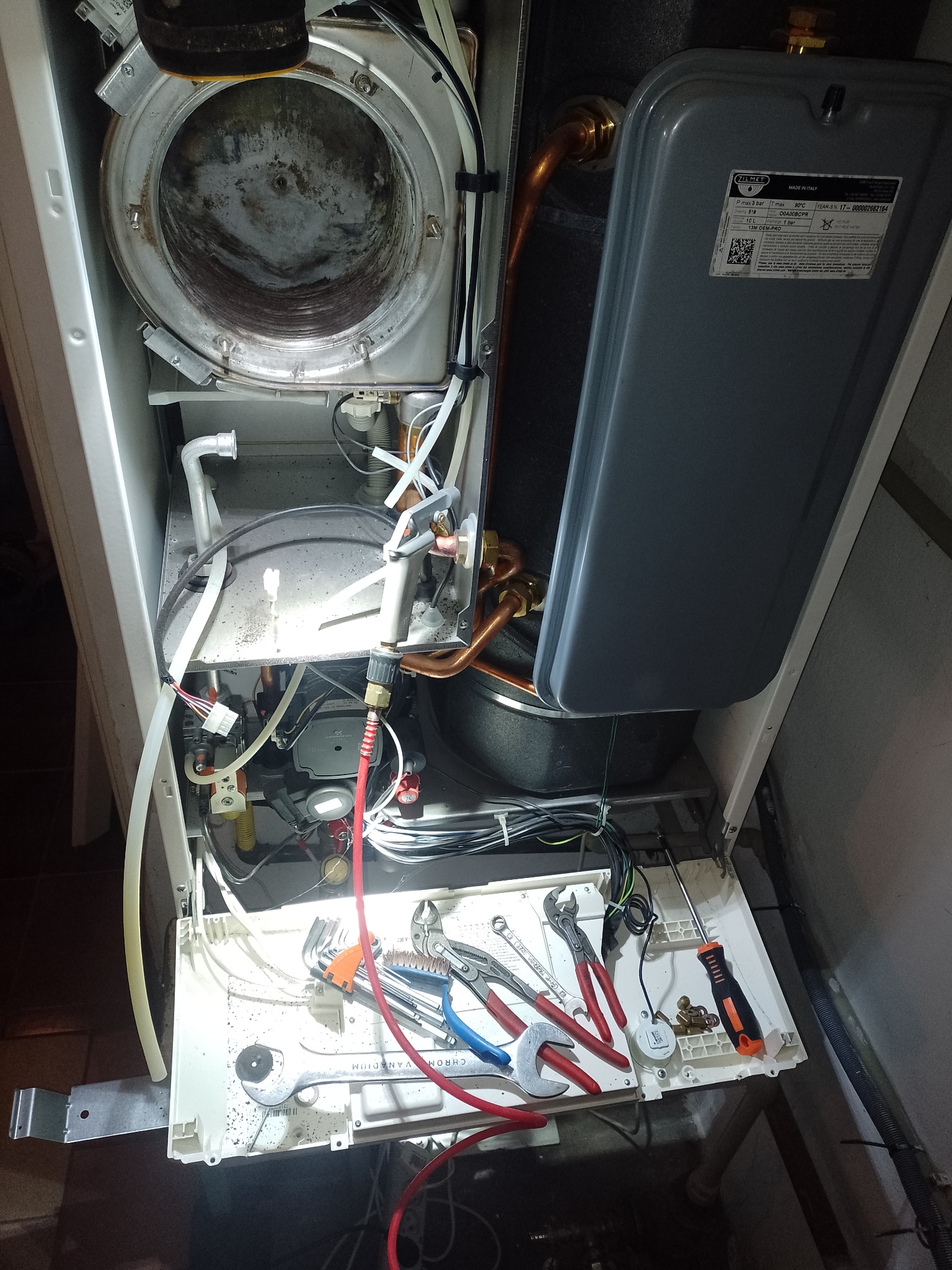 Wnętrze otwartego pieca gazowego z rozłożonymi narzędziami hydraulicznymi na dolnej obudowie, widoczne miedziane rury i elementy instalacji.