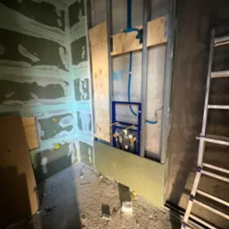 Wnętrze łazienki w trakcie remontu: ściany częściowo pokryte płytami gipsowo-kartonowymi i szpachlą, widoczne metalowe profile, drewniane elementy konstrukcyjne, instalacja wodno-kanalizacyjna...