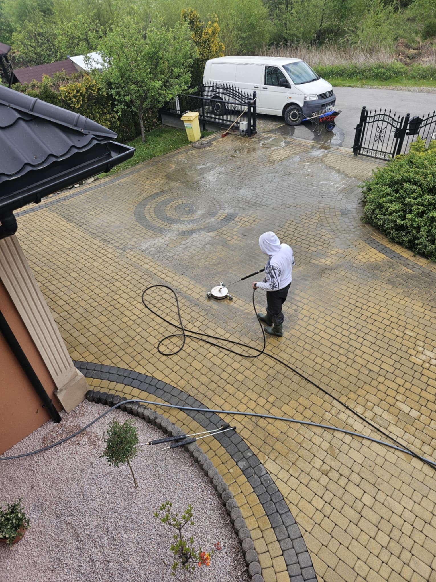 Czyszczenie kostki brukowej myjką ciśnieniową. Widoczna różnica w kolorze czyszczonej i nieczyszczonej powierzchni. Osoba w kombinezonie ochronnym podczas pracy.