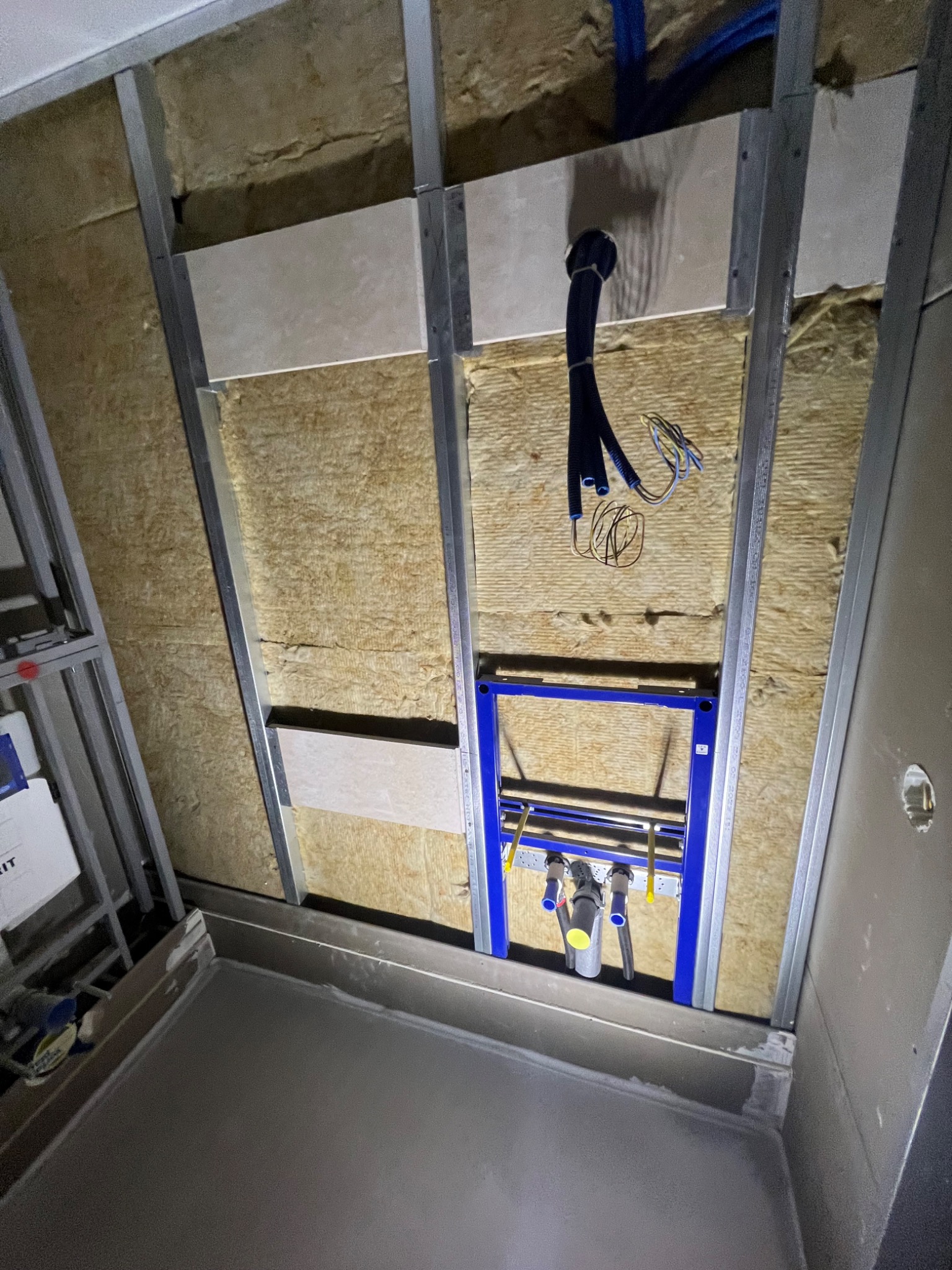 Wnętrze łazienki w trakcie prac: widoczna izolacja z wełny mineralnej, metalowa konstrukcja stelaża, fragment zabudowy płytami kartonowo-gipsowymi oraz niebieski stelaż podtynkowy WC z podłączonymi...