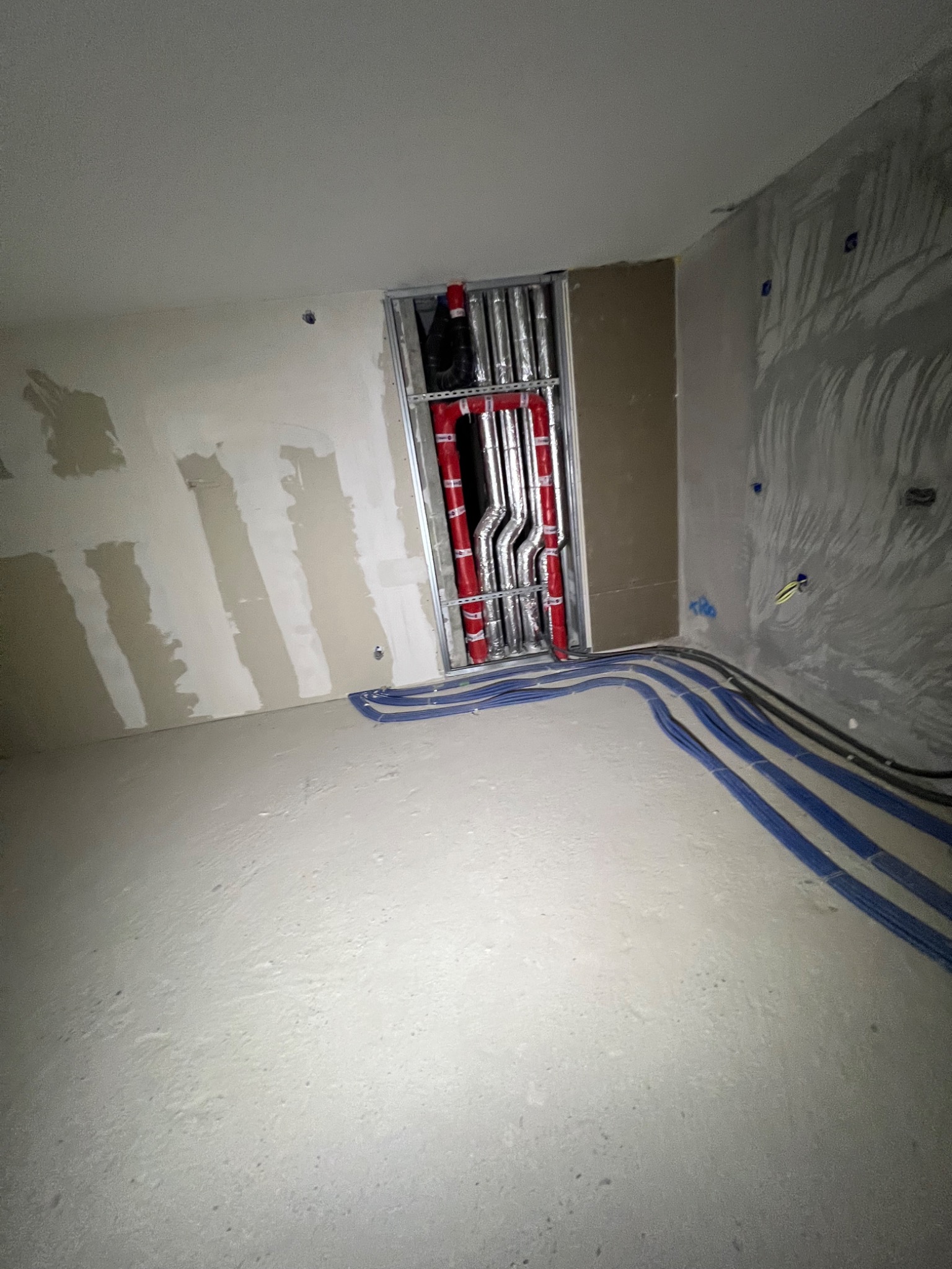 Wnętrze w trakcie prac wykończeniowych: ściany po szpachlowaniu, widoczne rury instalacji grzewczej w metalowej ramie i niebieskie przewody elektryczne na podłodze.