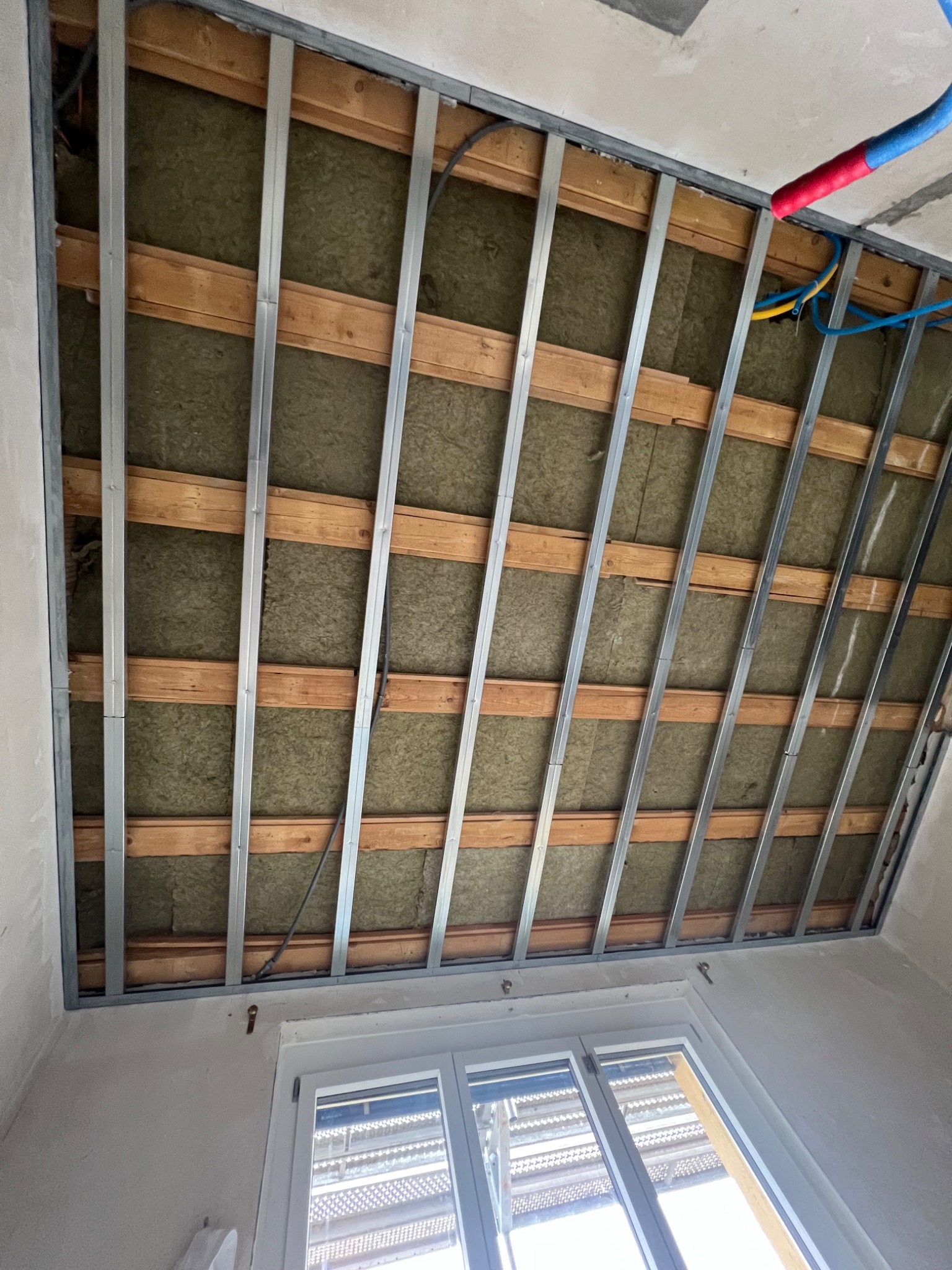 Szkielet sufitu podwieszanego z widoczną izolacją z wełny mineralnej, metalowe profile i drewniane belki, instalacja elektryczna w tle, okno w dolnej części kadru.