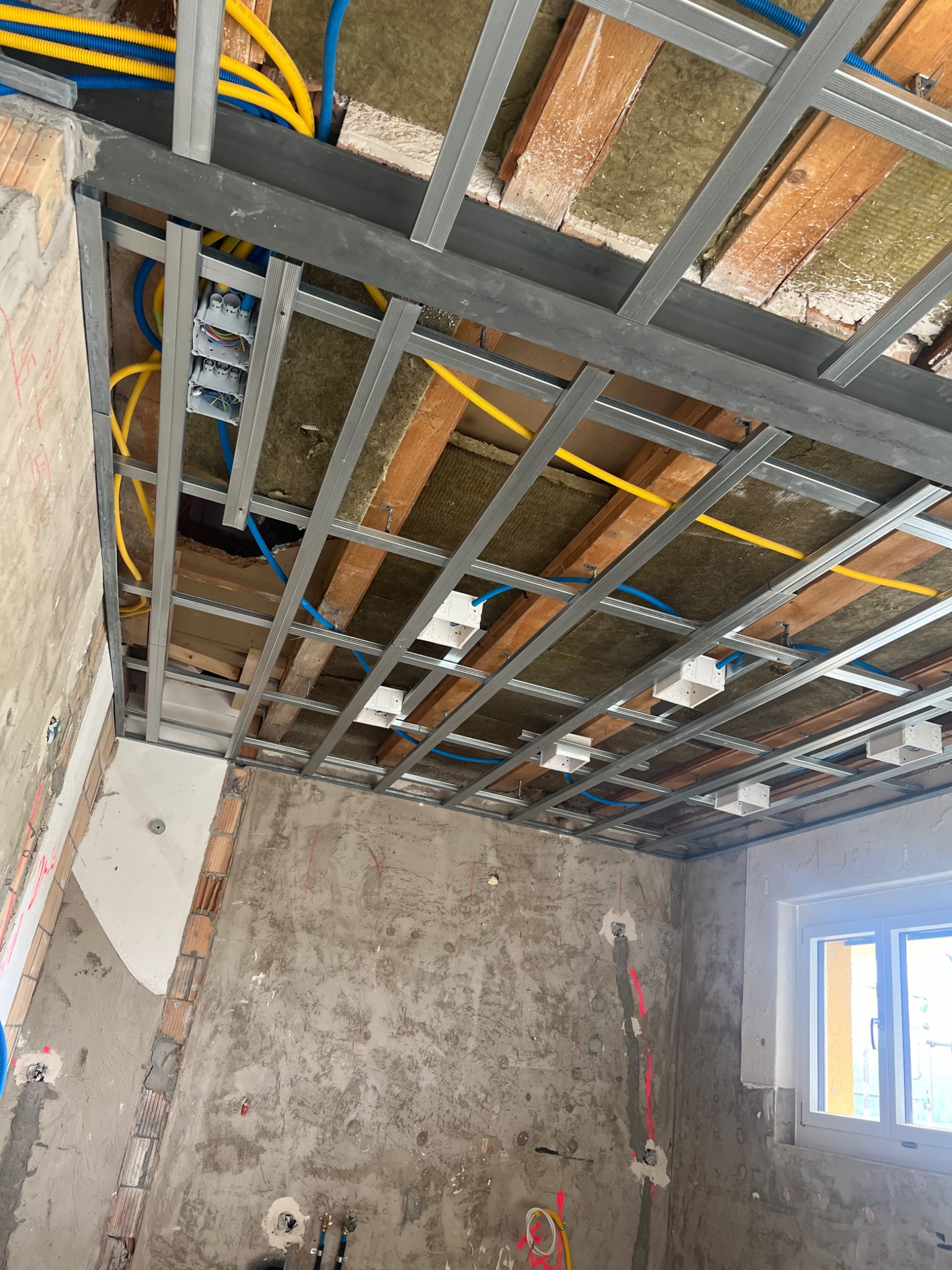 Montaż metalowego stelażu sufitu podwieszanego z widoczną izolacją termiczną, instalacją elektryczną i hydrauliczną w surowym wnętrzu z oknem.