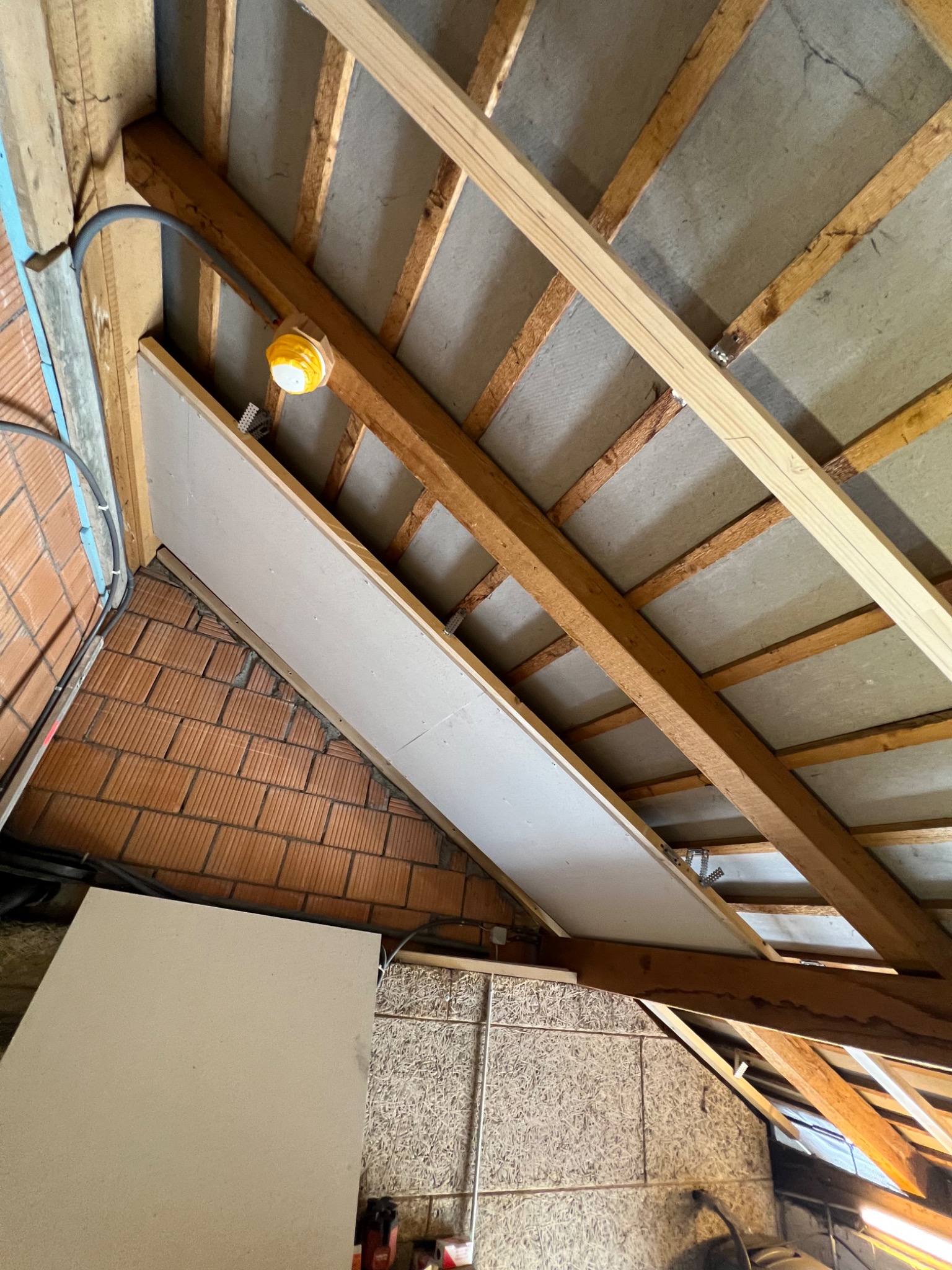 Poddasze z widoczną konstrukcją drewnianą, izolacją z szarej wełny mineralnej i częściowo zamontowaną płytą gipsowo-kartonową, oświetlone żółtą lampą roboczą.