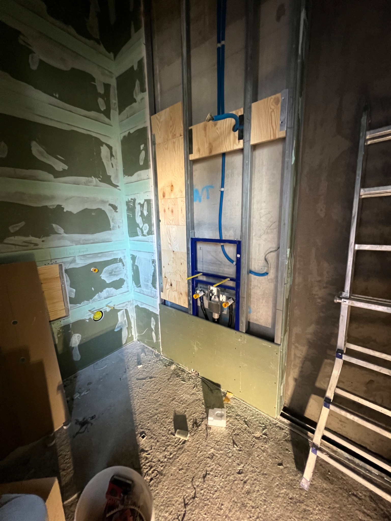 Wnętrze łazienki w trakcie remontu: ściany częściowo pokryte płytami gipsowo-kartonowymi i szpachlą, widoczne metalowe profile, drewniane elementy konstrukcyjne, instalacja wodno-kanalizacyjna...