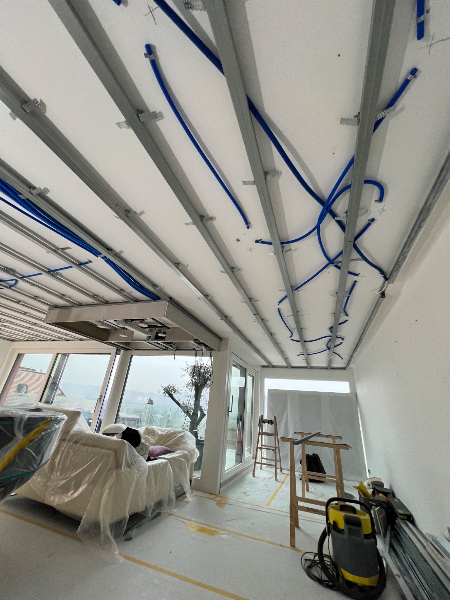 Montaż sufitu podwieszanego z widocznymi profilami metalowymi, przewodami wentylacyjnymi i oświetleniem, meble zabezpieczone folią, drabina i odkurzacz na podłodze.