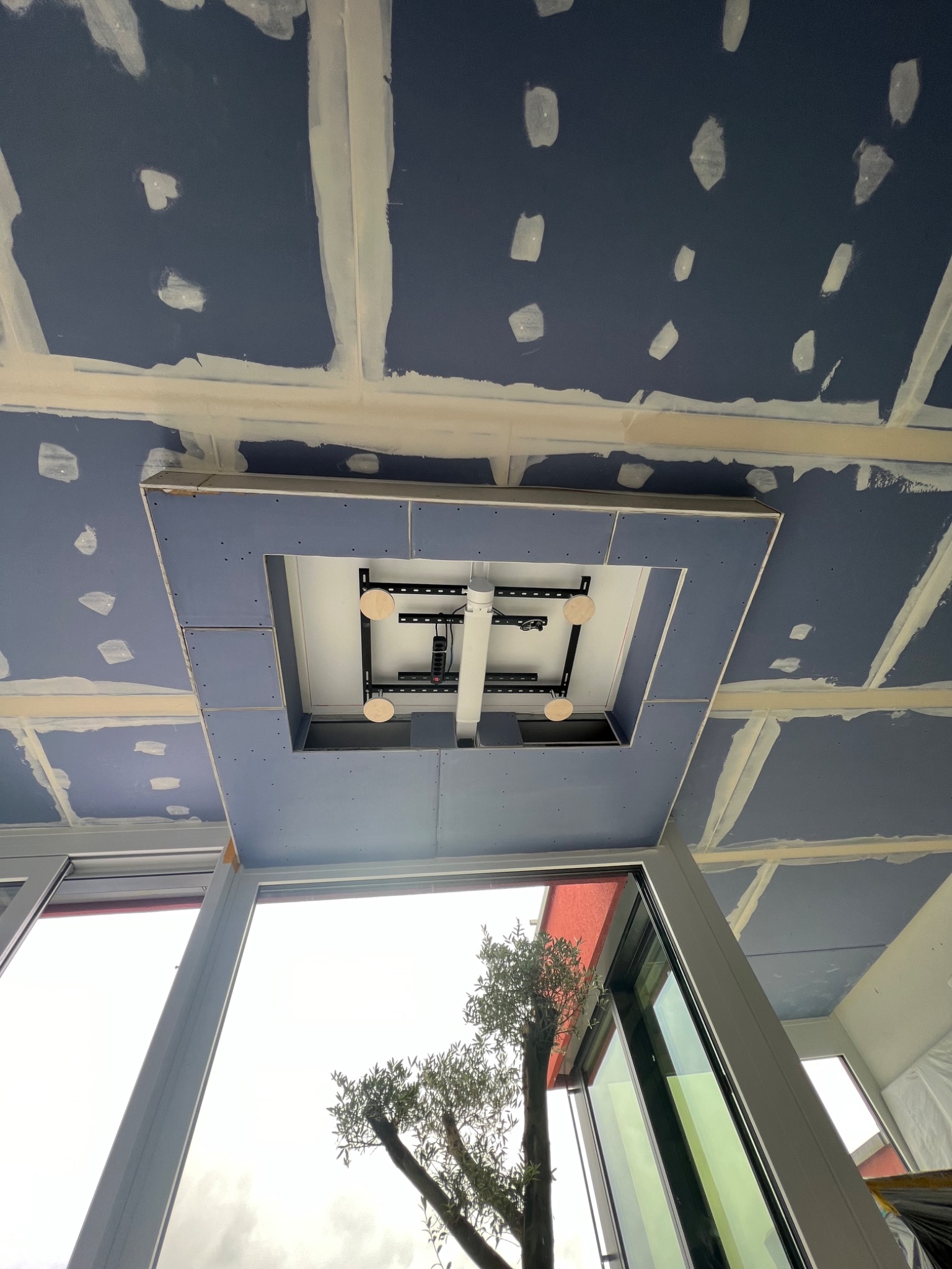 Niebieski sufit podwieszany z widoczną konstrukcją metalową i oświetleniem, częściowo zaszpachlowany, z widokiem na okno i drzewo na zewnątrz.