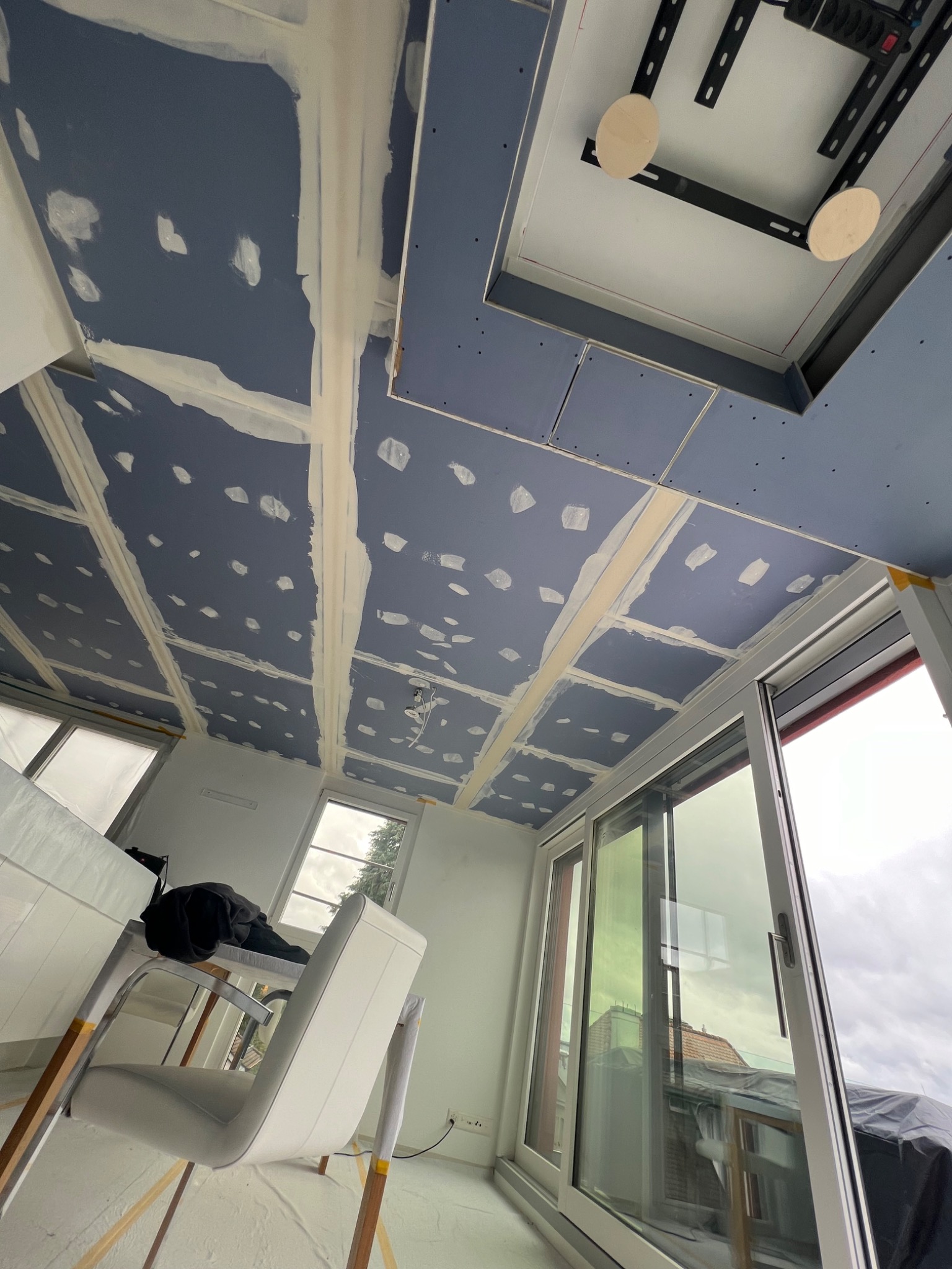 Płyty gipsowo-kartonowe na suficie w trakcie szpachlowania, widoczne łączenia wypełnione masą szpachlową, narożniki oklejone taśmą malarską, okno balkonowe przesuwne z widokiem na pochmurne niebo.