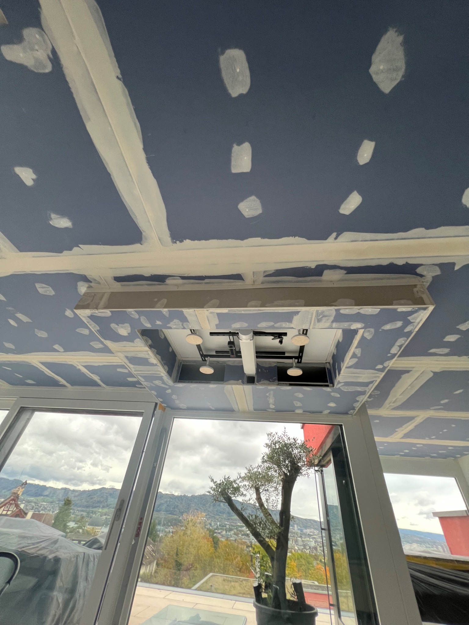 Płyty gipsowo-kartonowe na suficie w trakcie szpachlowania, widoczne łączenia wypełnione masą, przygotowanie do malowania, w tle widok przez okno na miasto i góry.